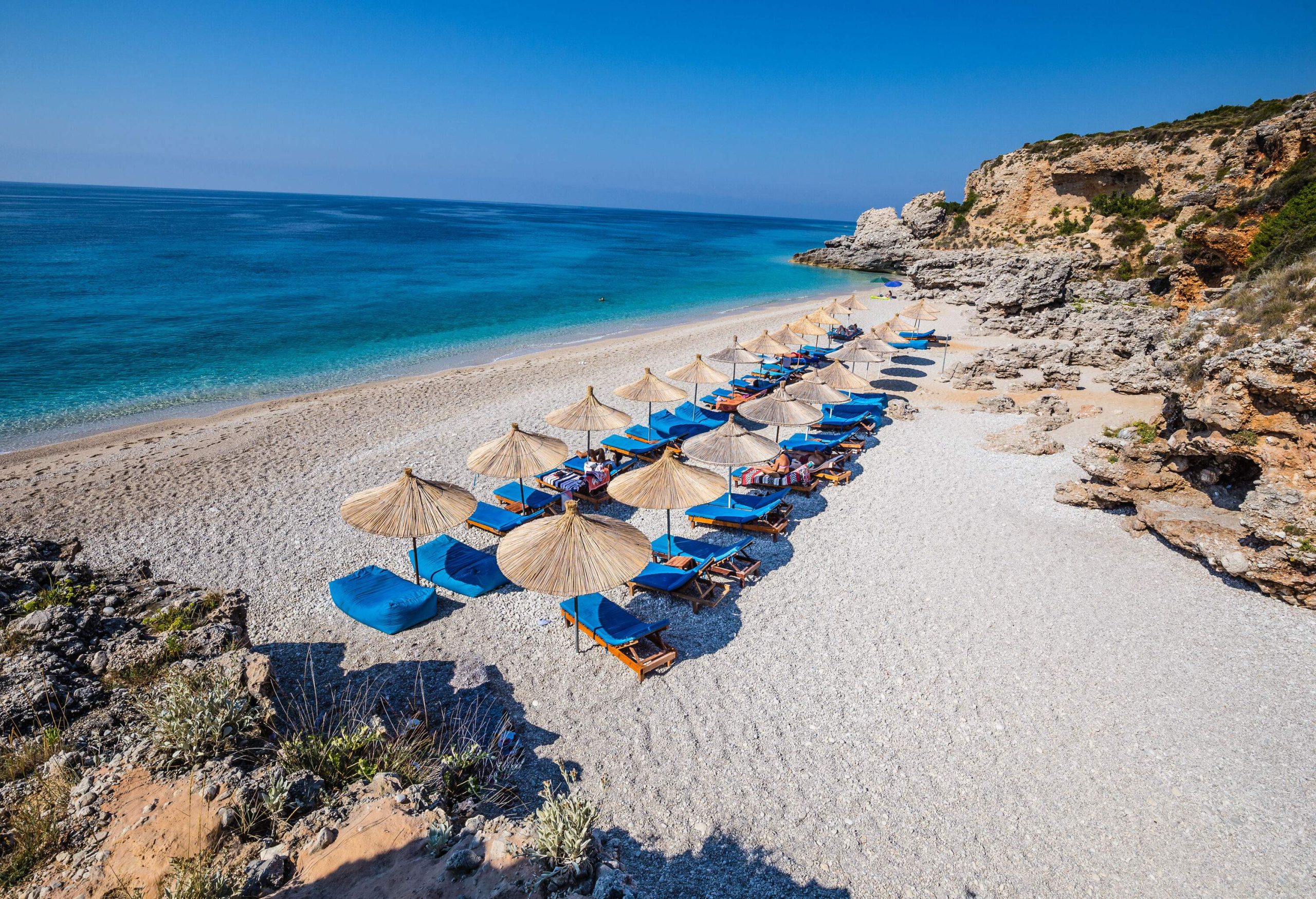 Two rows of parasols and sun loungers line a narrow beach, nestled between the glistening sea and rugged rocky surroundings.