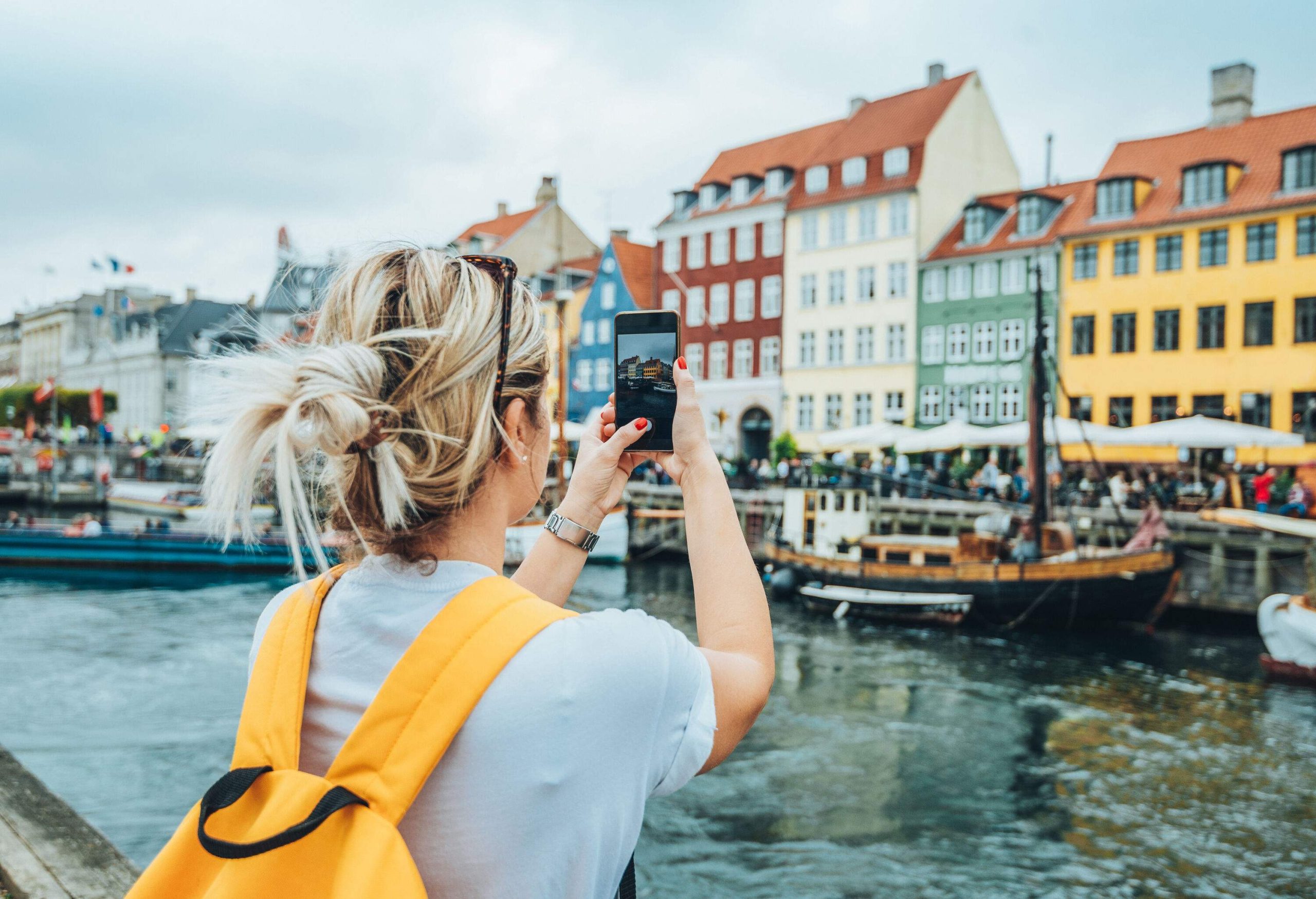 A woman using her smartphone to take a photo of the colourful buildings along a harbour.