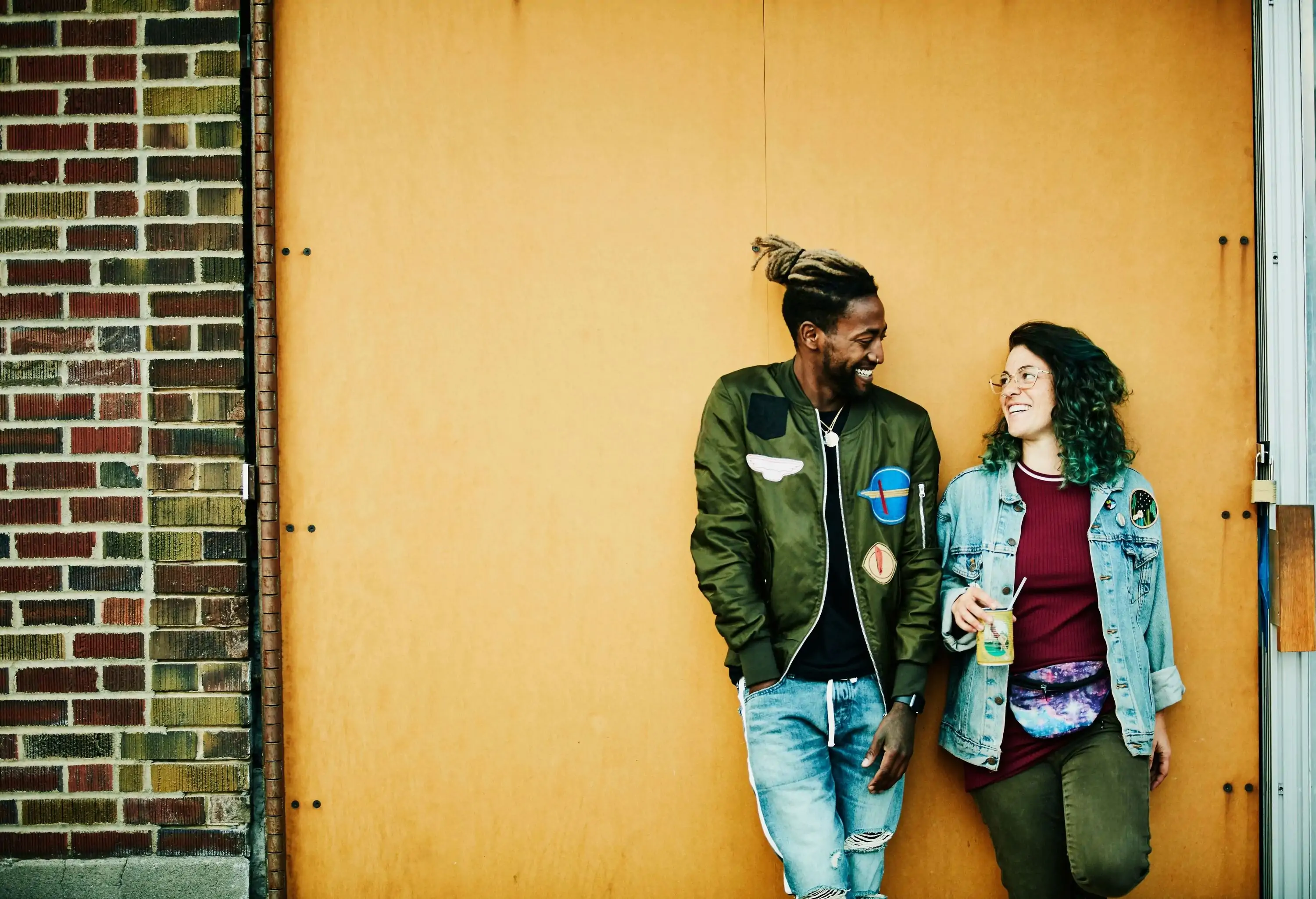 A young couple leaning against a wall, looking at each other with big smiles on their faces.