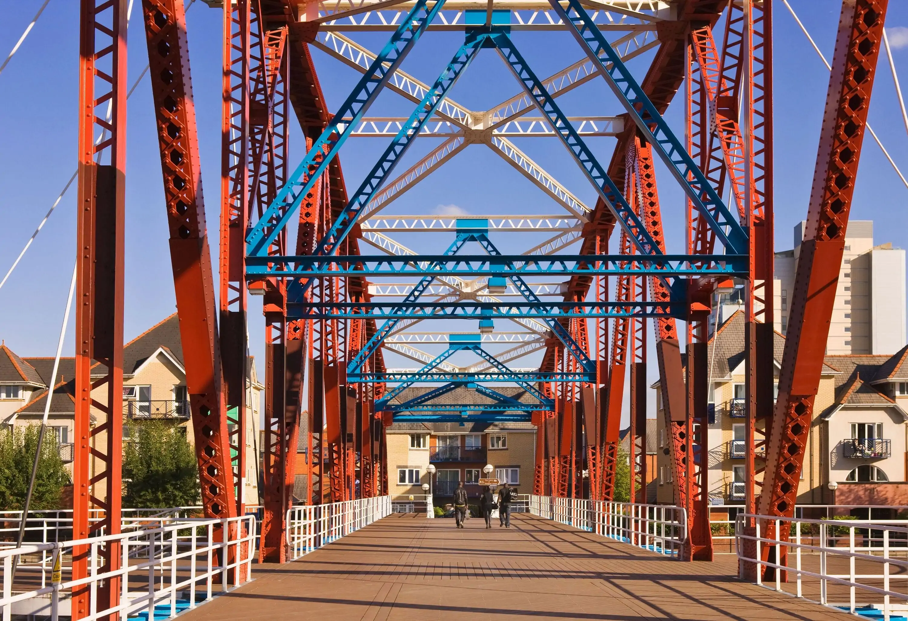 A group of people nearing the end of a pedestrian overpass adorned with colourful metalwork and leading to a residential area.