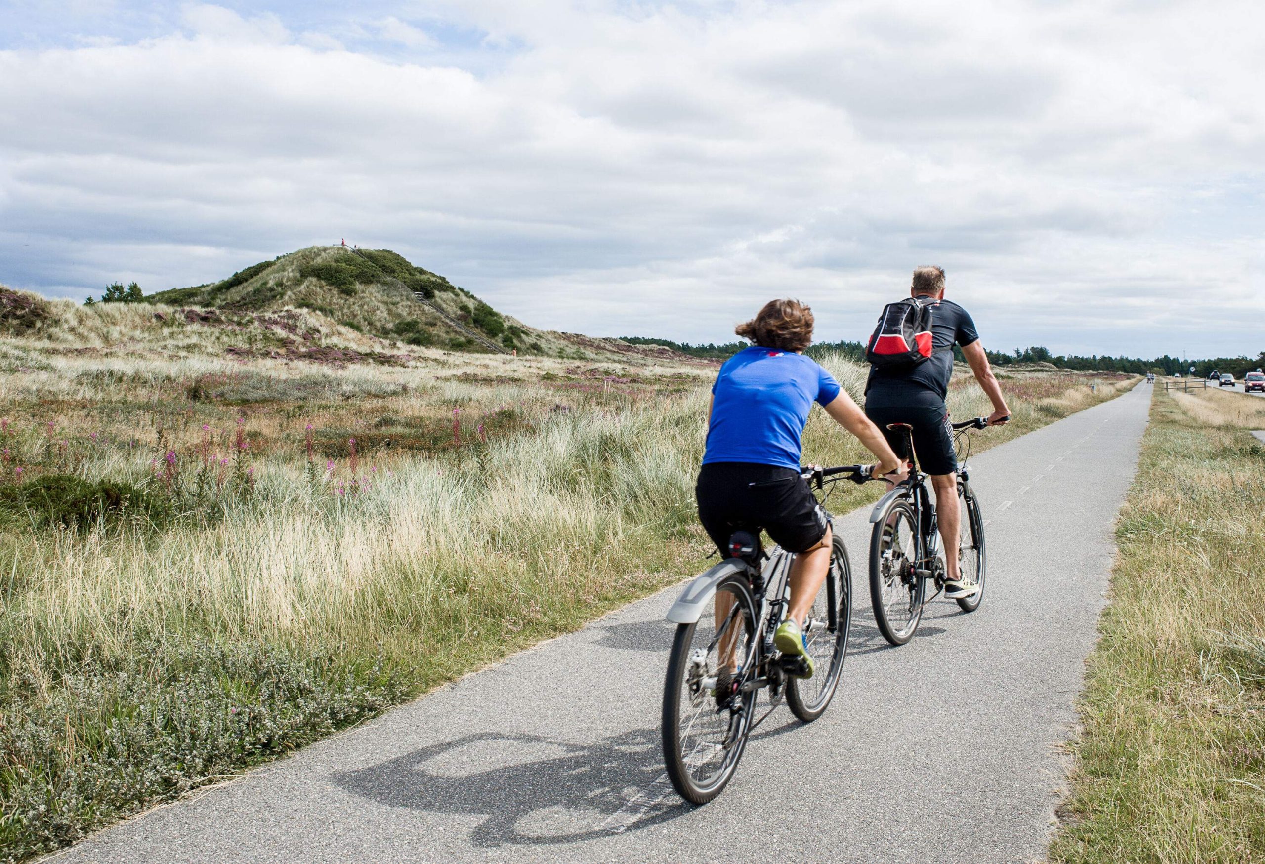Two people pedal on the paved road where the grassland continues on the side of the main road.