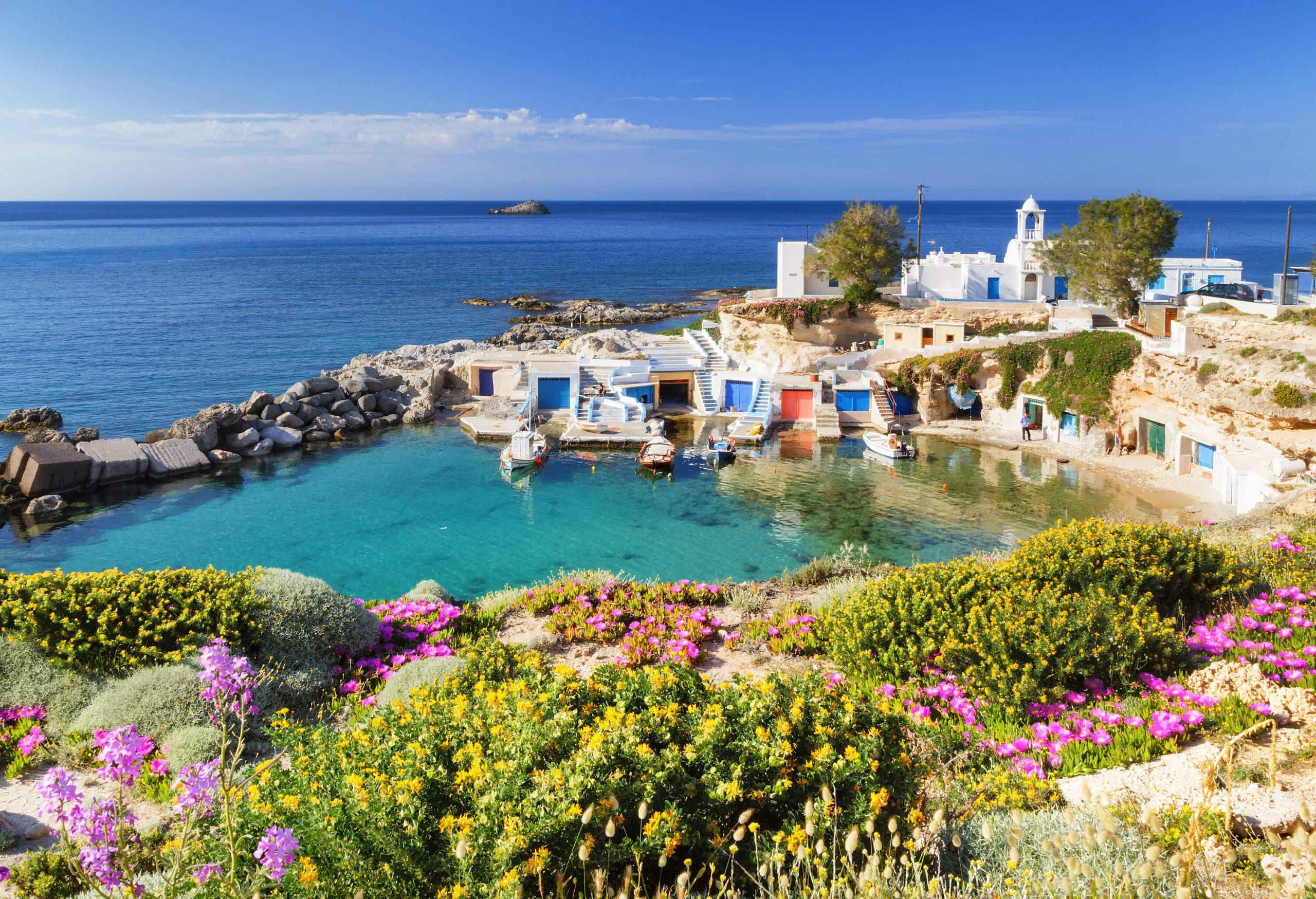 A settlement tucked within the rocky terrain surrounding a body of water, with a blue sea visible in the background and colourful plants in the foreground.
