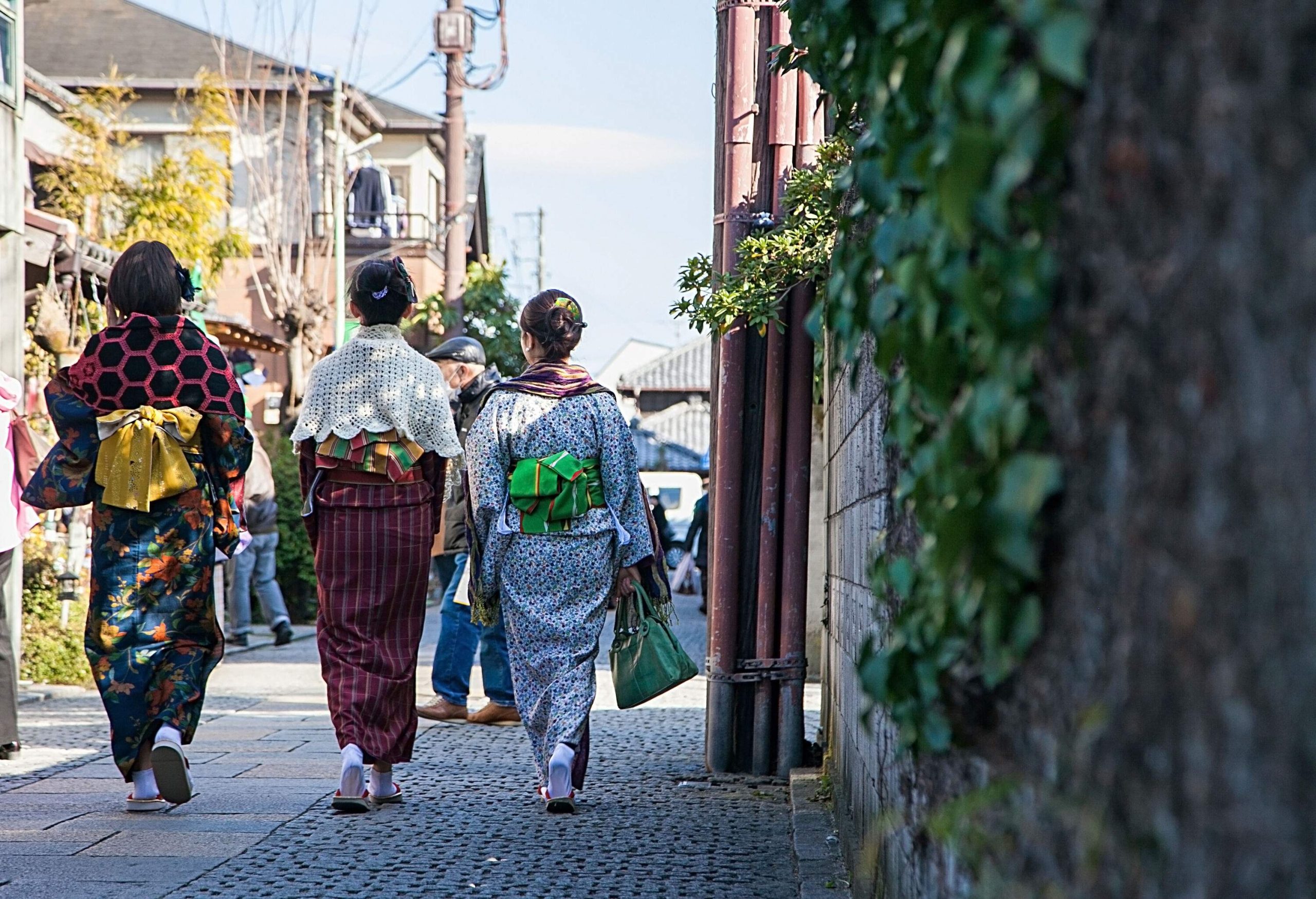 Three women wear colourful kimonos as they walk on a street in a residential neighbourhood.