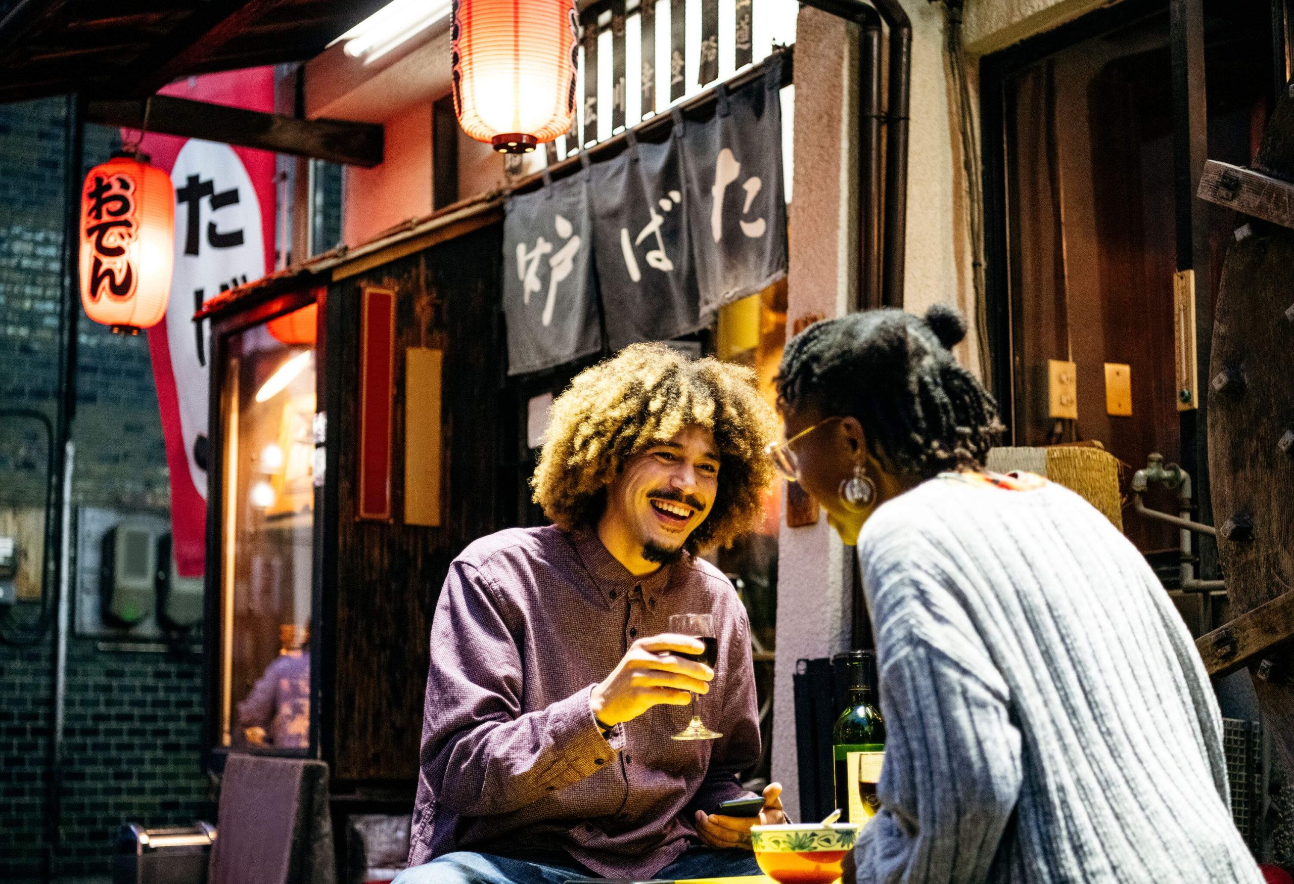 Young man with Afro hair laughing with friend and sitting outside Japanese Izakaya