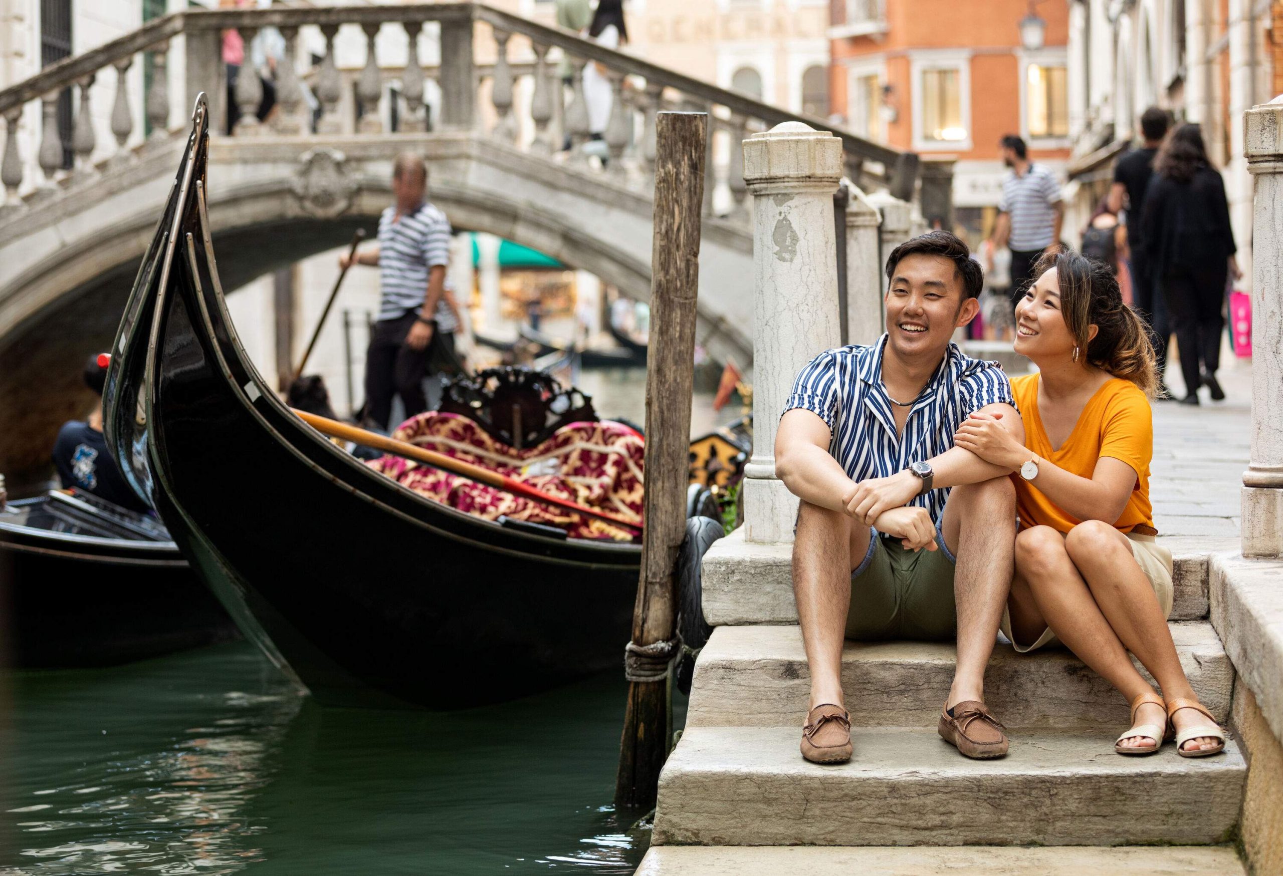 A happy couple sits on the stairs of a water canal with sailing gondolas under an arch bridge.