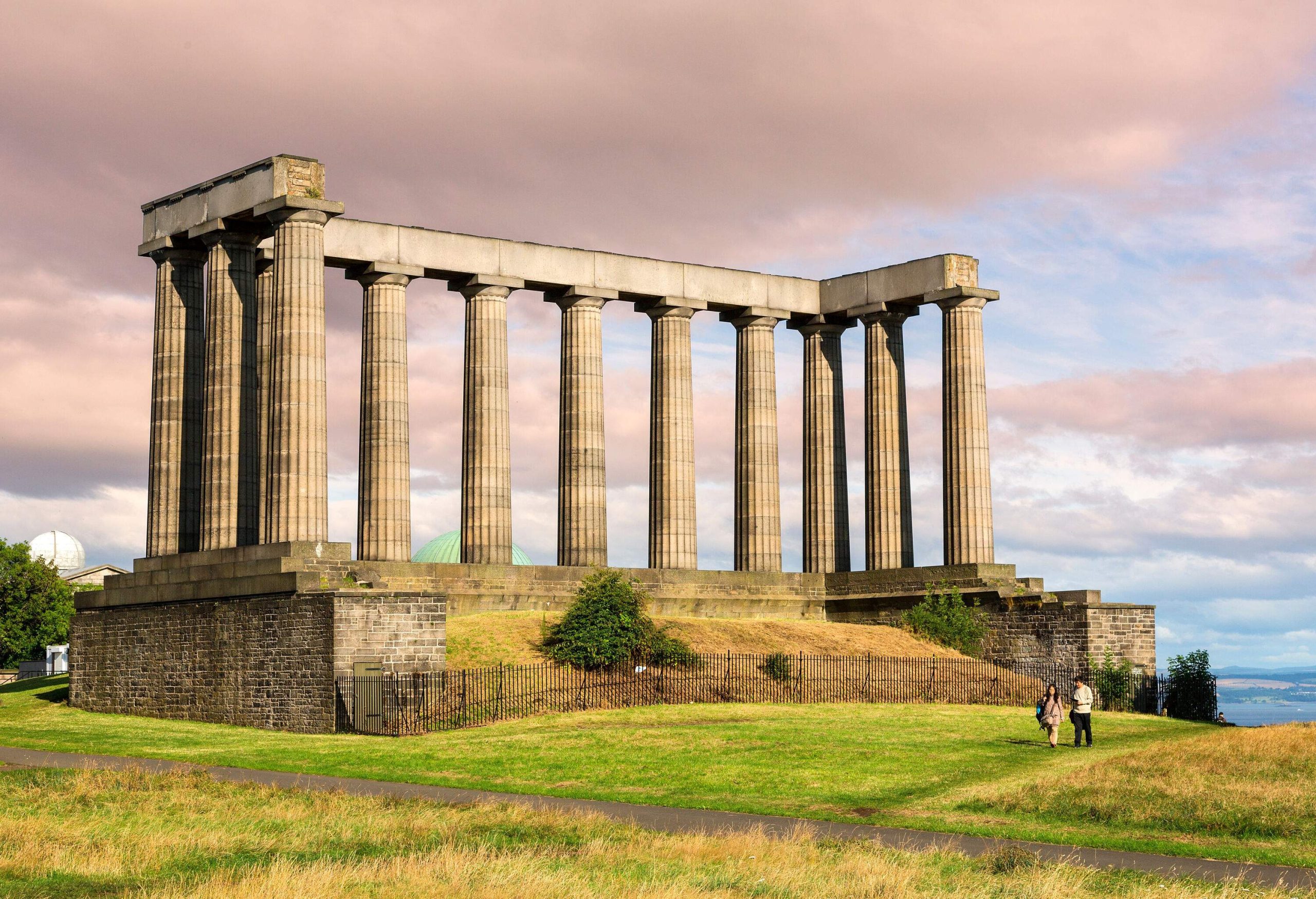 Two people stroll across a grass field next to a pillared monument.