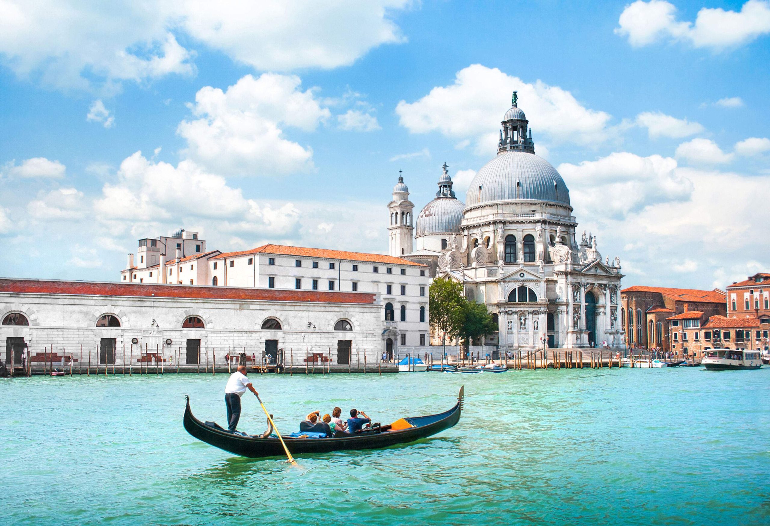 A gondola carrying people on Canal Grande with the Basilica di Santa Maria della Salute visible in the background.