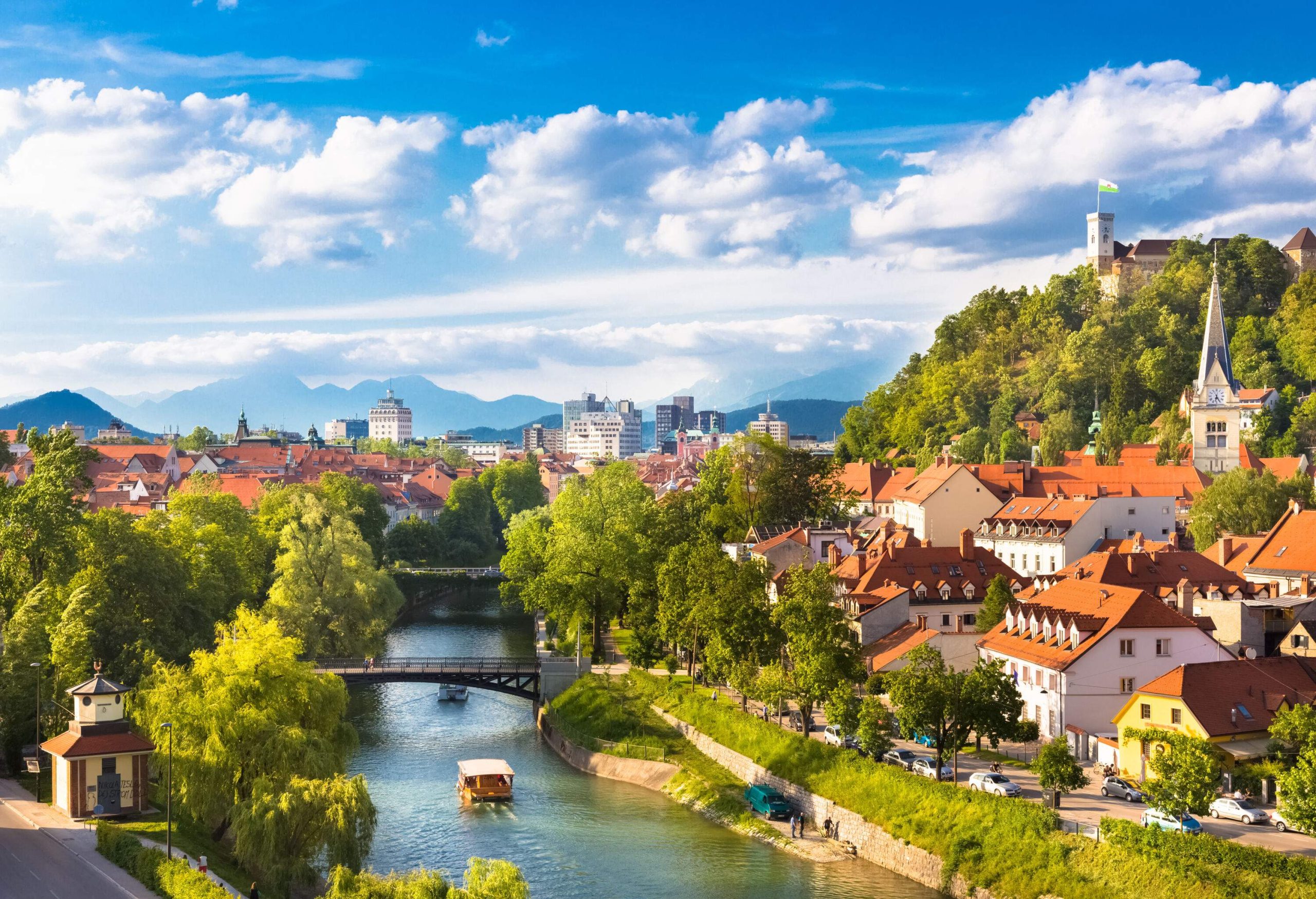 A boat cruising down a river with historic structures and beautiful trees covering the banks and hilltops.