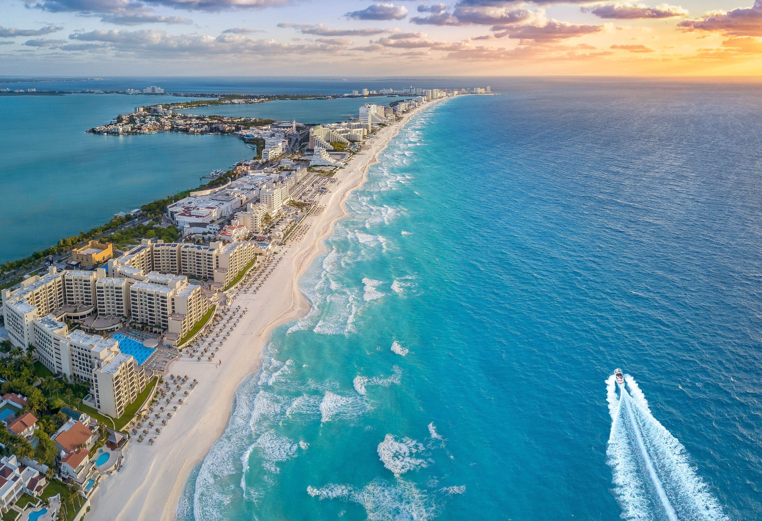 Aerial view of a white sand beach and a coastal city on a lush island surrounded by the blue sea against the sunset sky.