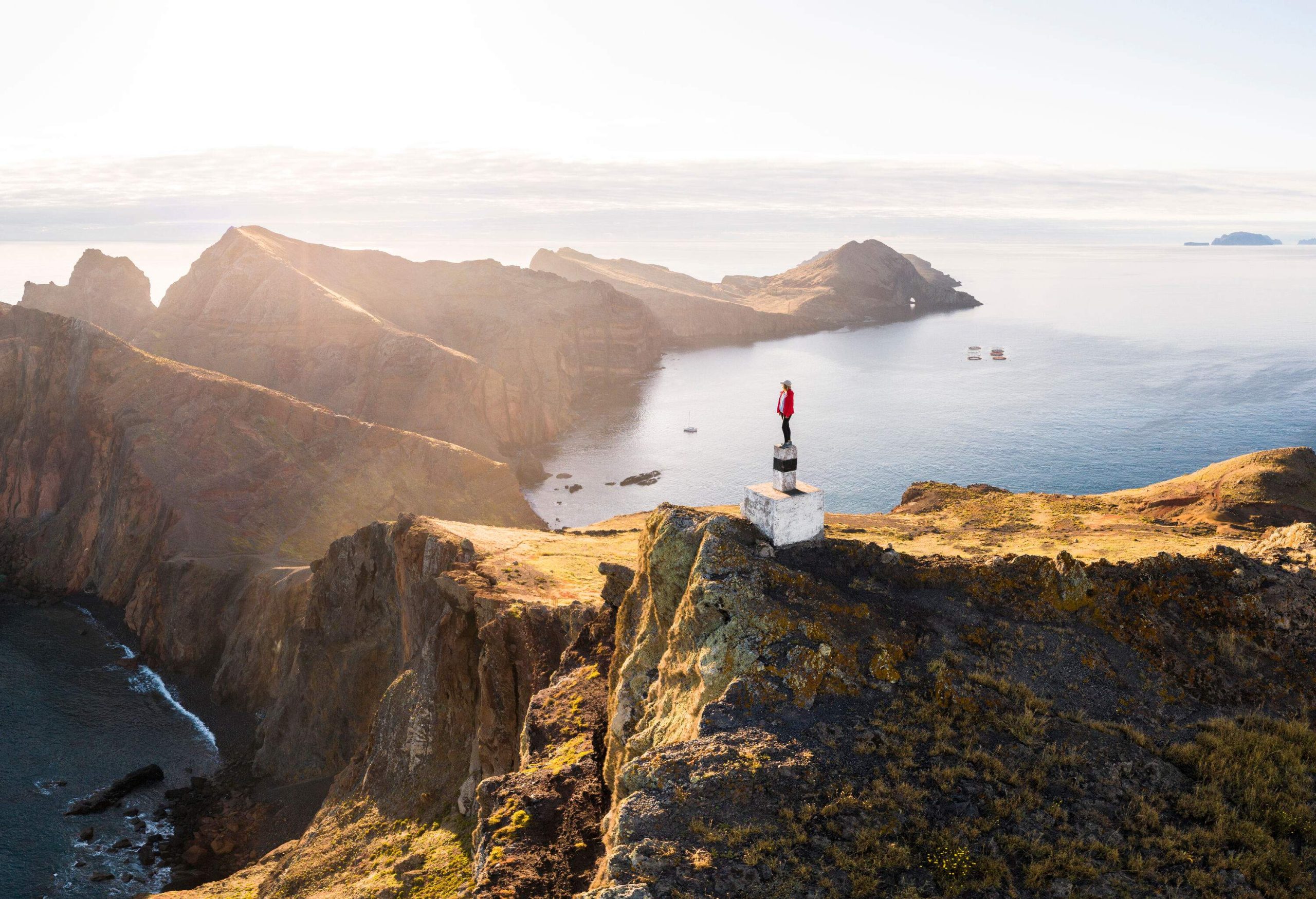 A person stands on a white concrete slab of a summit with views of the surrounding cliffs, mountain ridges, and enormous ocean.