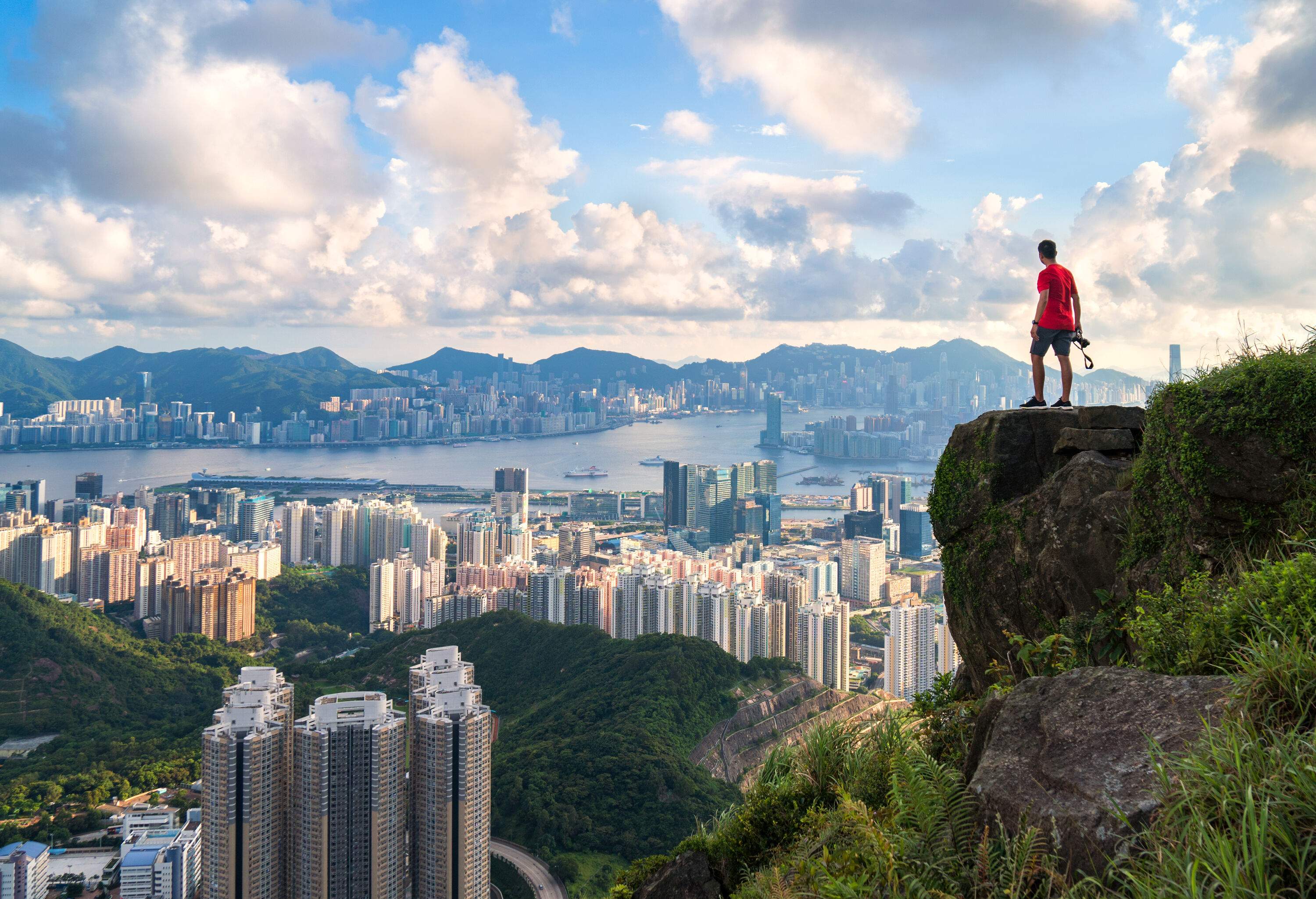 A male photographer stands at the mountaintop edge overlooking the sprawling cityscape against a dramatic sky.