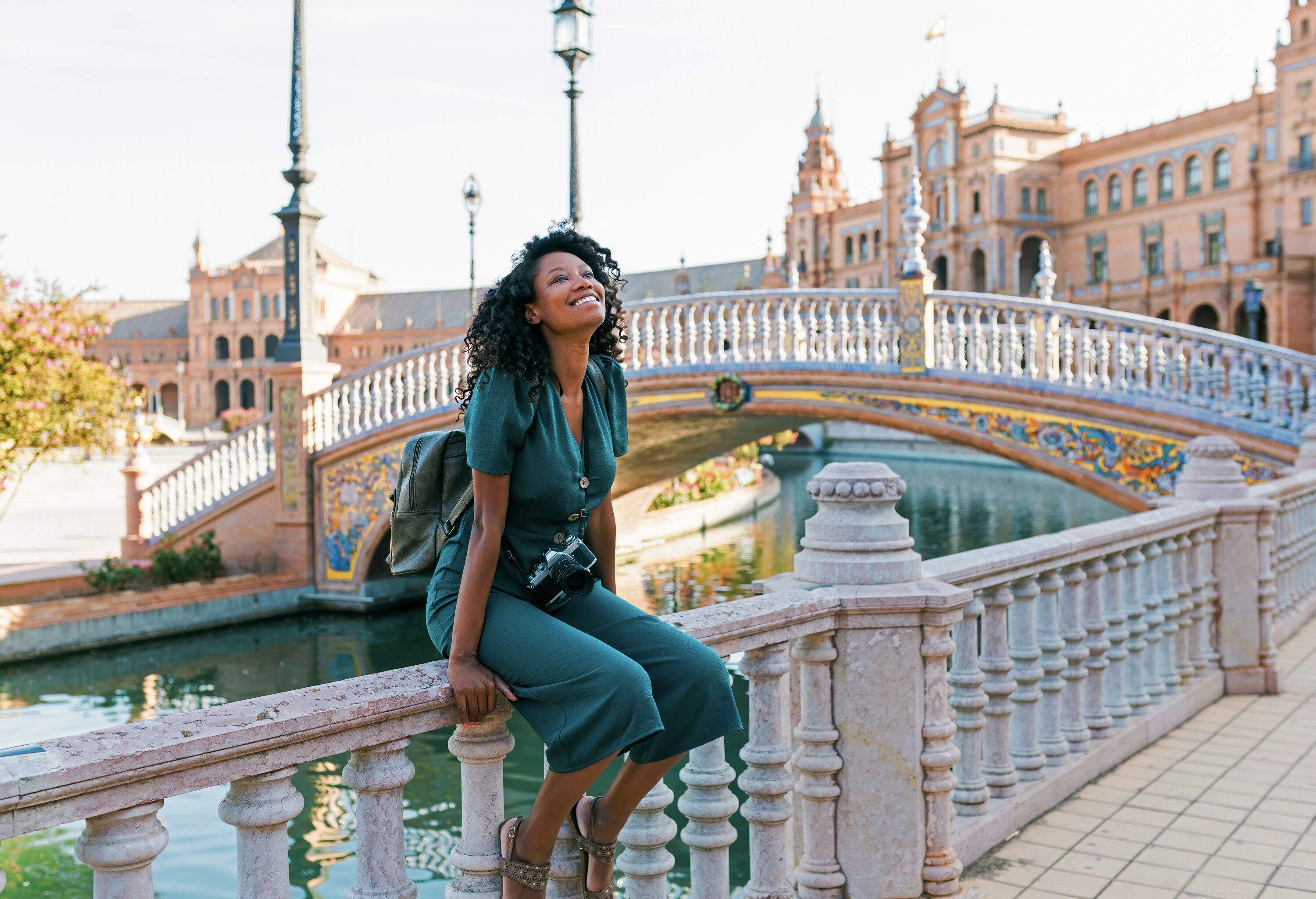 A woman in casual attire smiles as she sits on the railings of the sidewalk next to a bridge.