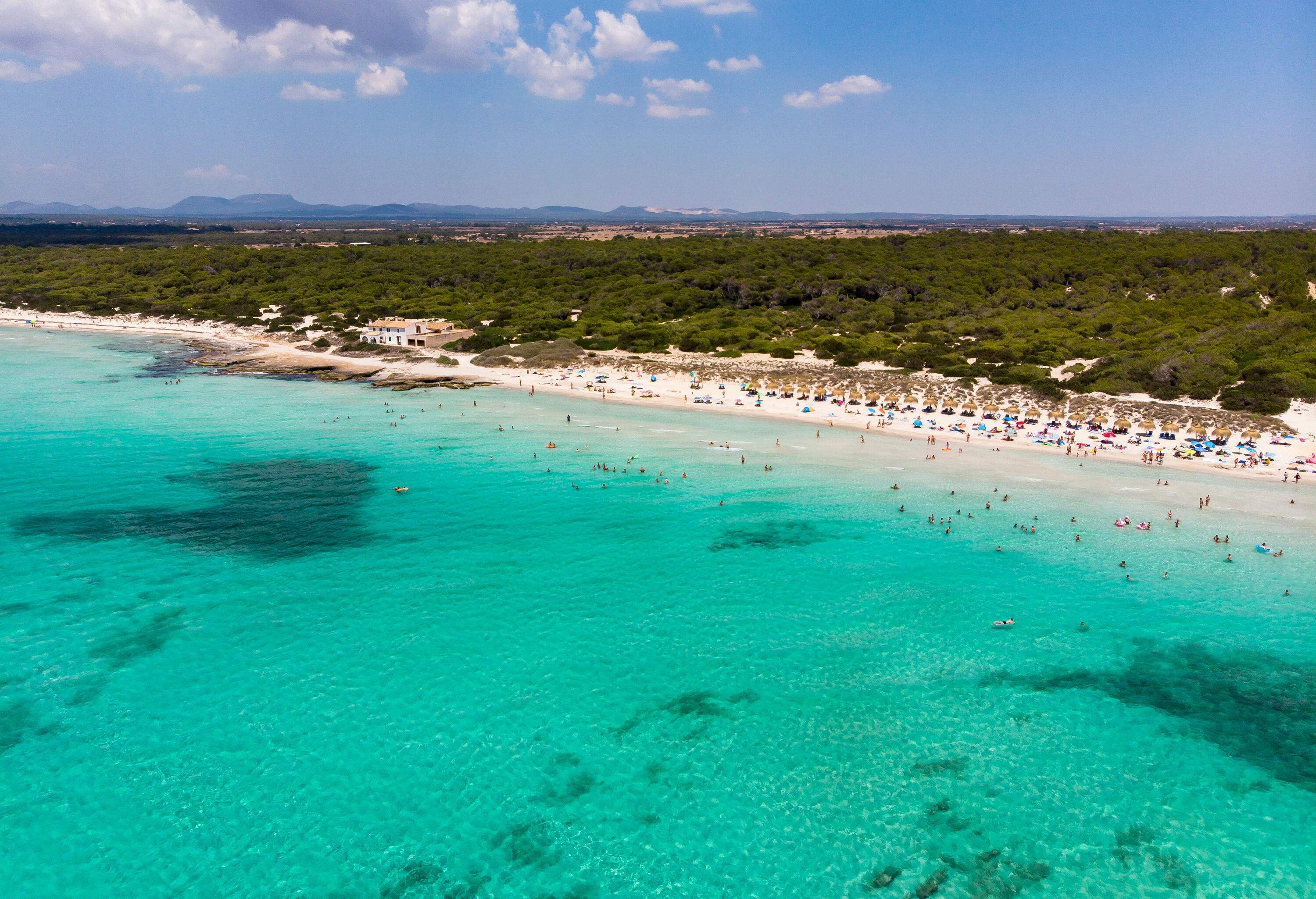 Aerial view of the turquoise sea and a lush island with a white sand shore lined with colourful parasols and sunbeds.