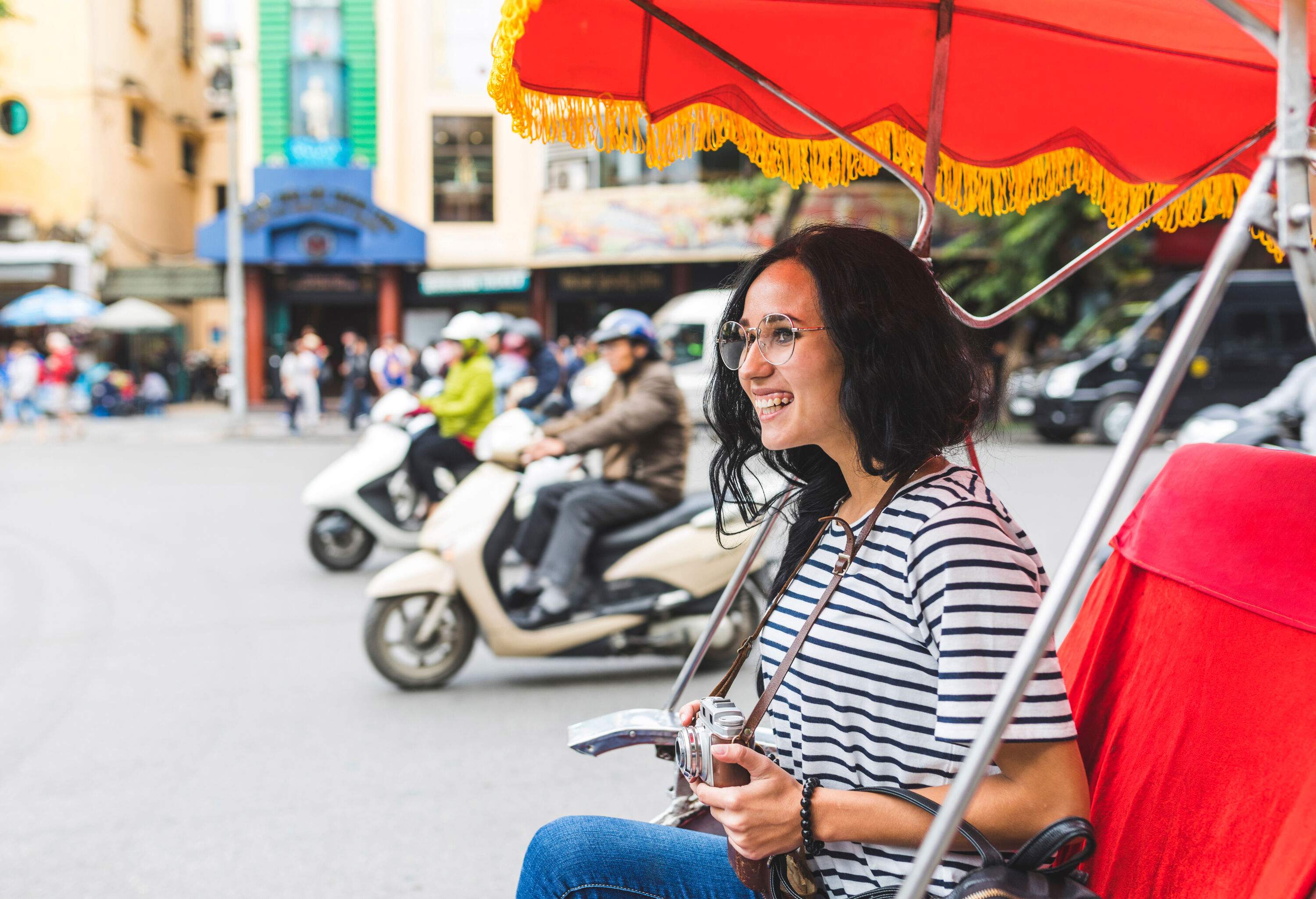 A woman in casual clothes smiles as she rides a rickshaw while holding a camera.