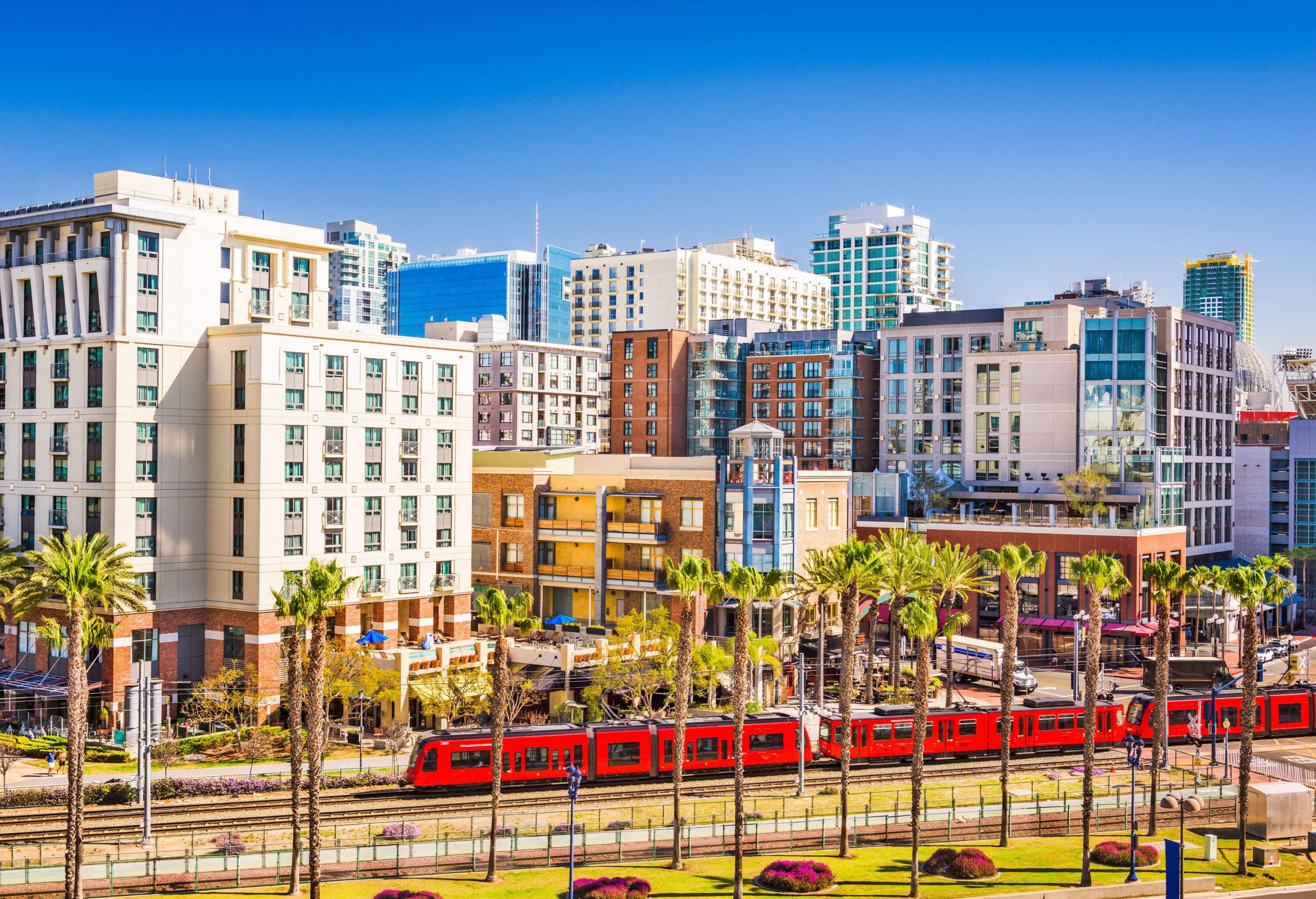 A red train running on a railway between a landscaped lawn with tall trees and a cluster of urban-style structures.