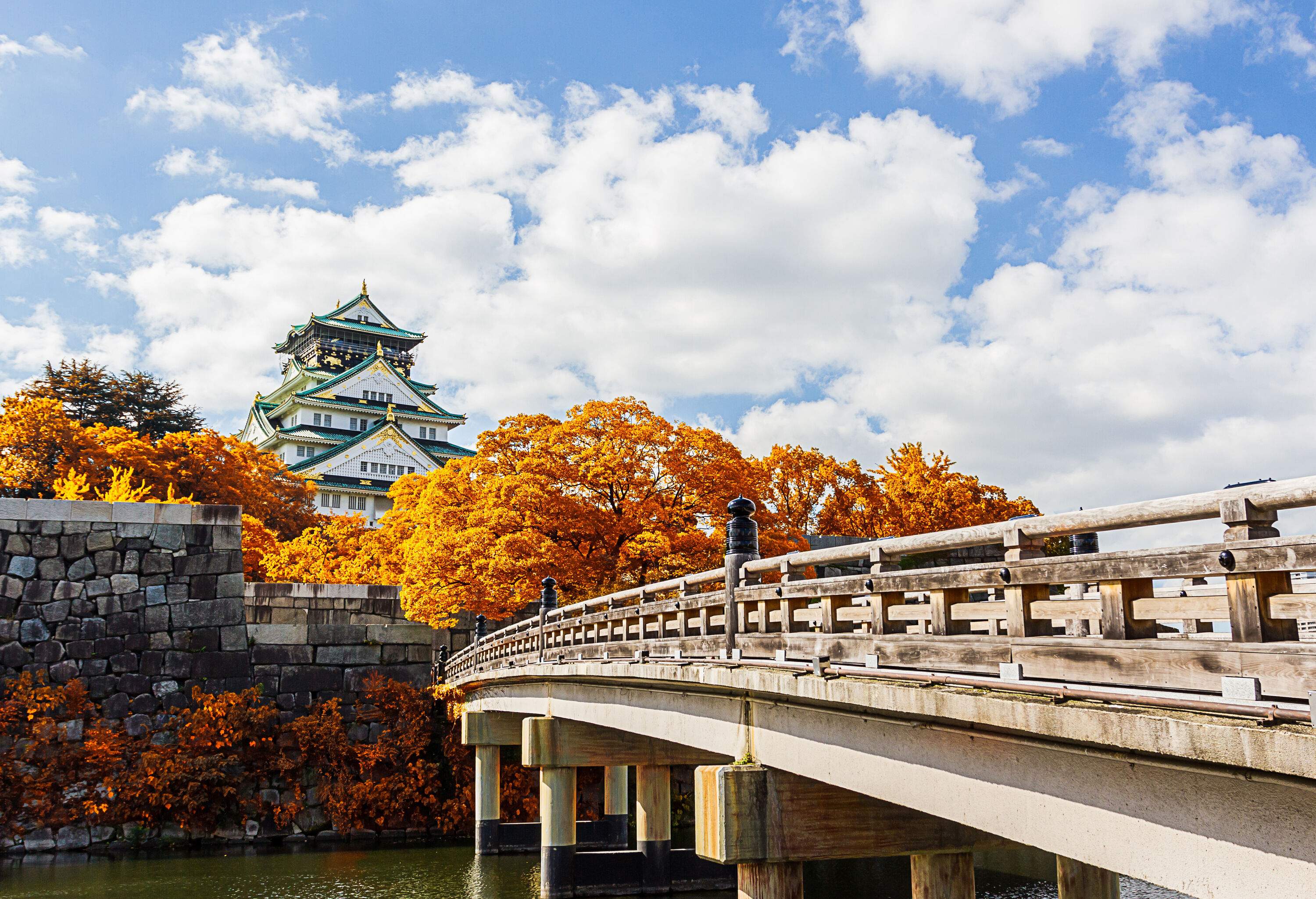 A wooden bridge over a moat with a view of the castle emerging from golden trees.
