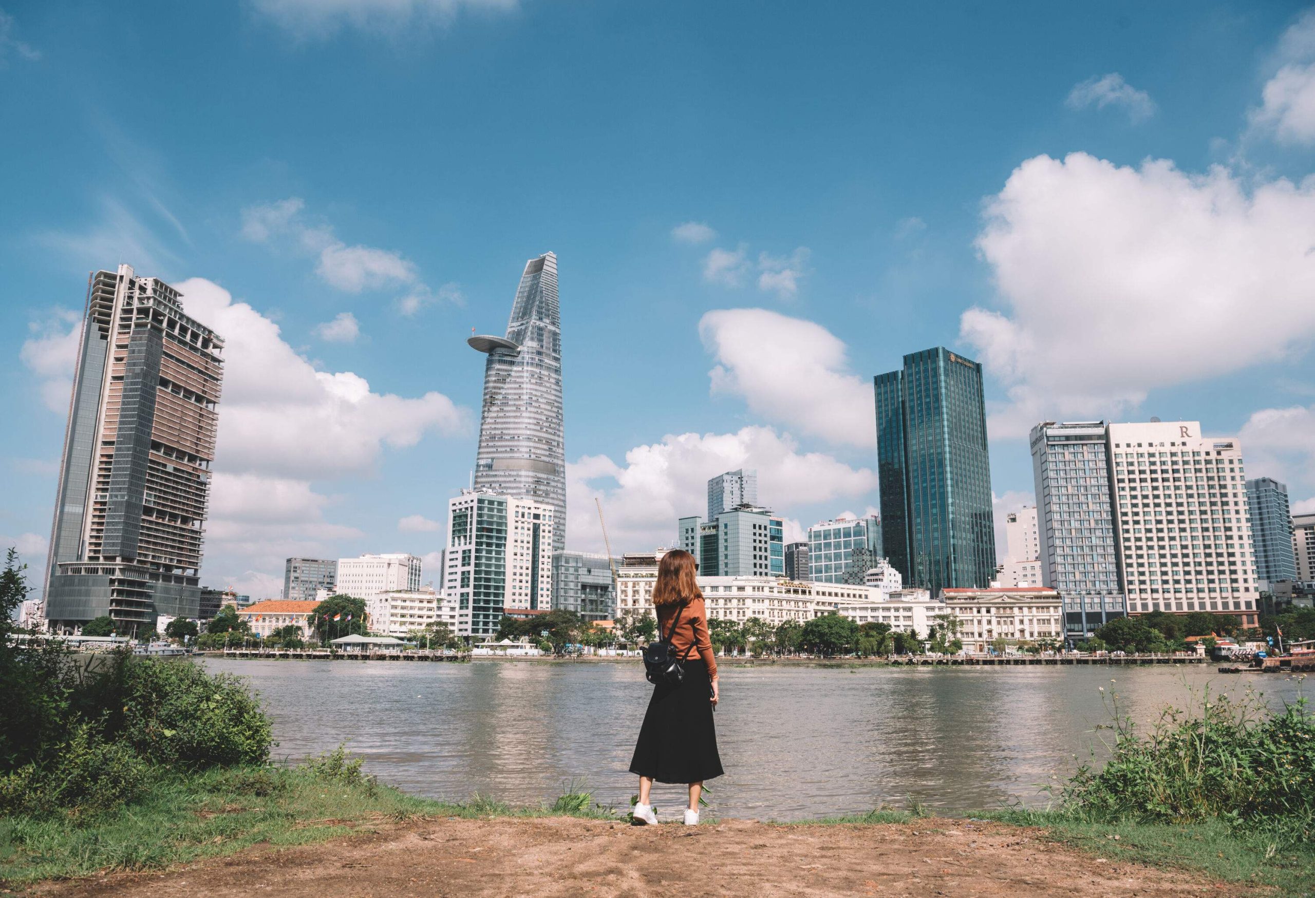 A woman in a brown sweater and black skirt stands on a riverbank against the backdrop of the city skyline.