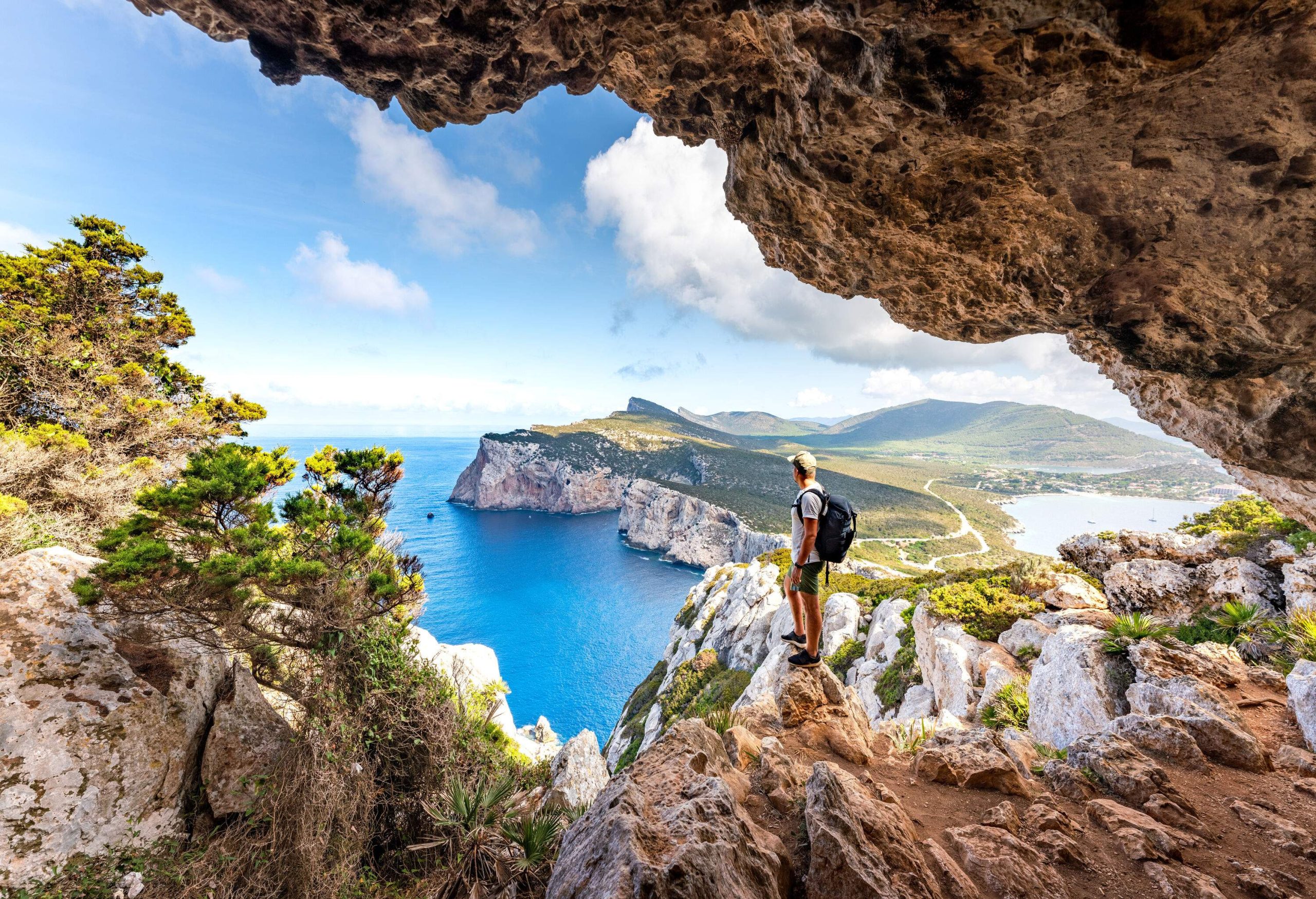 A person wearing a black backpack standing on the cliff of a rocky cave looking at the sea and a rocky island beneath.