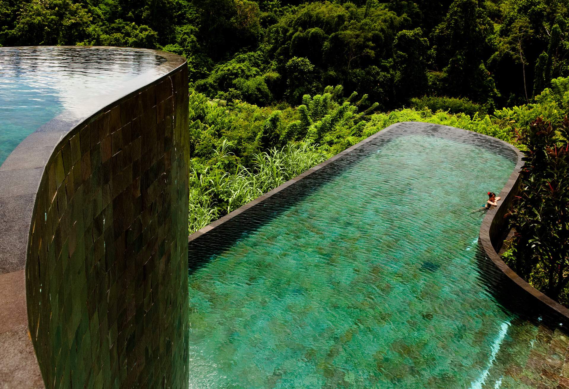 A woman leaning on the edge of a big swimming pool surrounded by dense woodland.