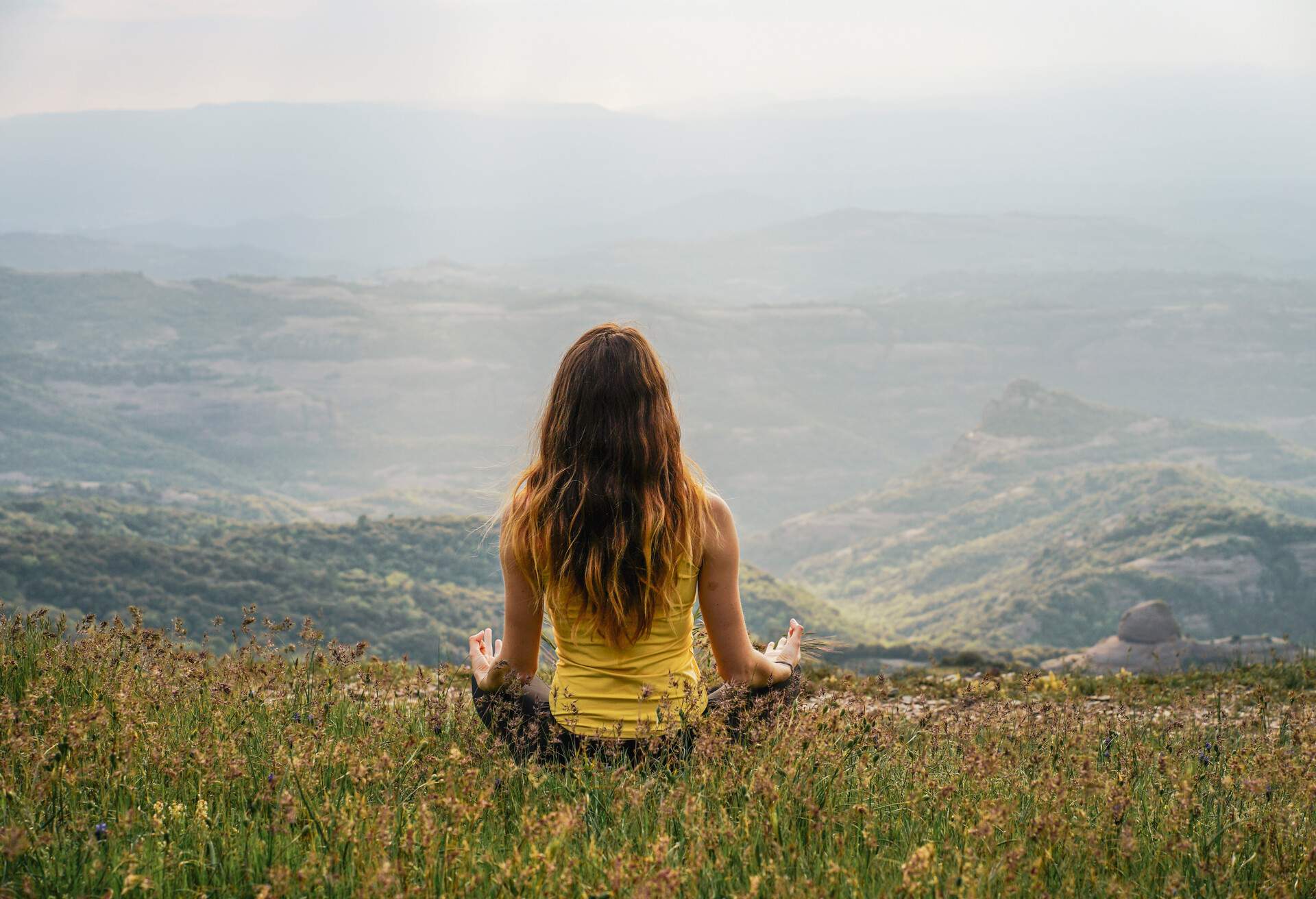 A woman in yellow shirt sit in a meditative pose on a hill with overlooking views of the mountains.