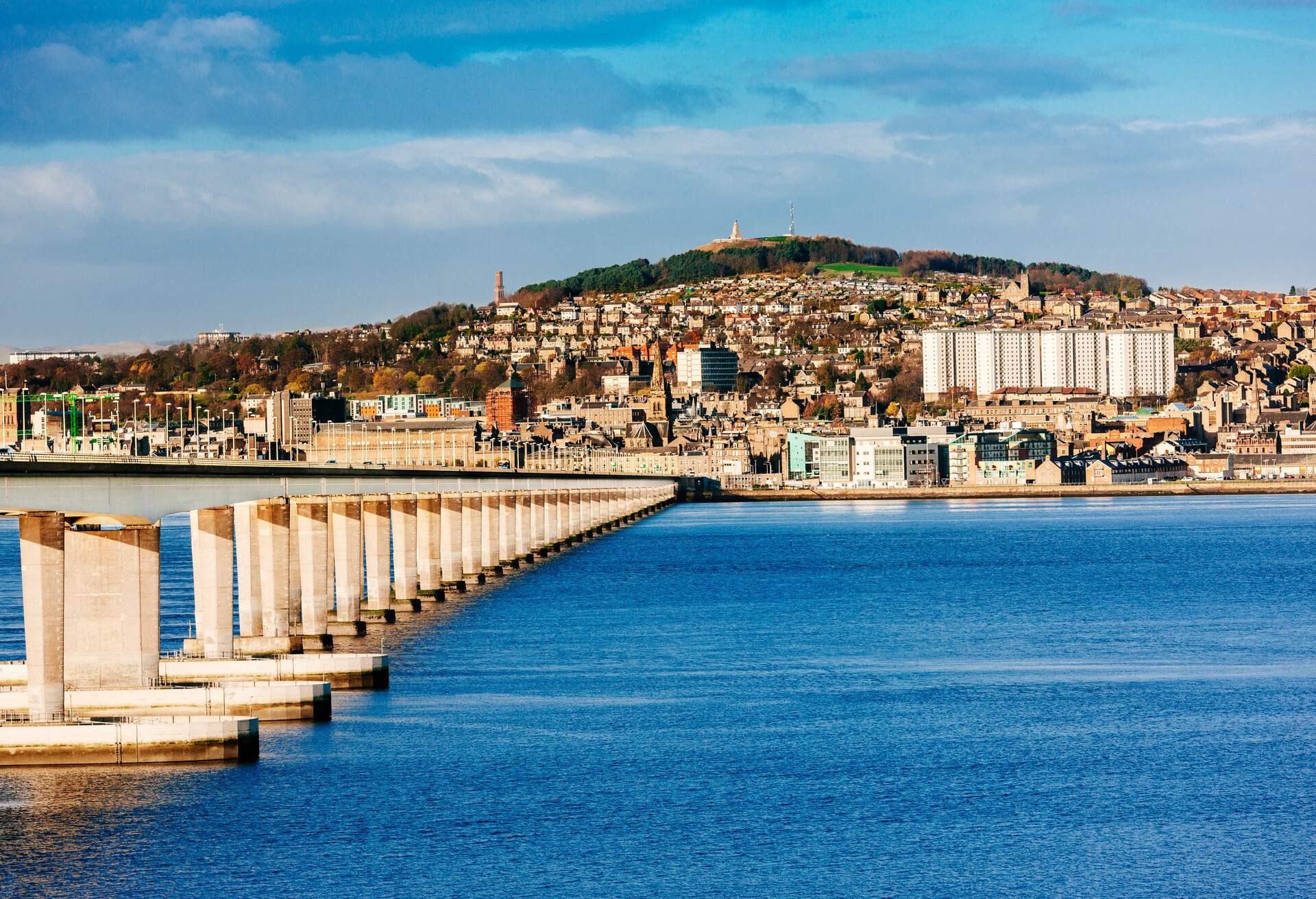 A road bridge supported by multiple spans with a hillside town in the background.