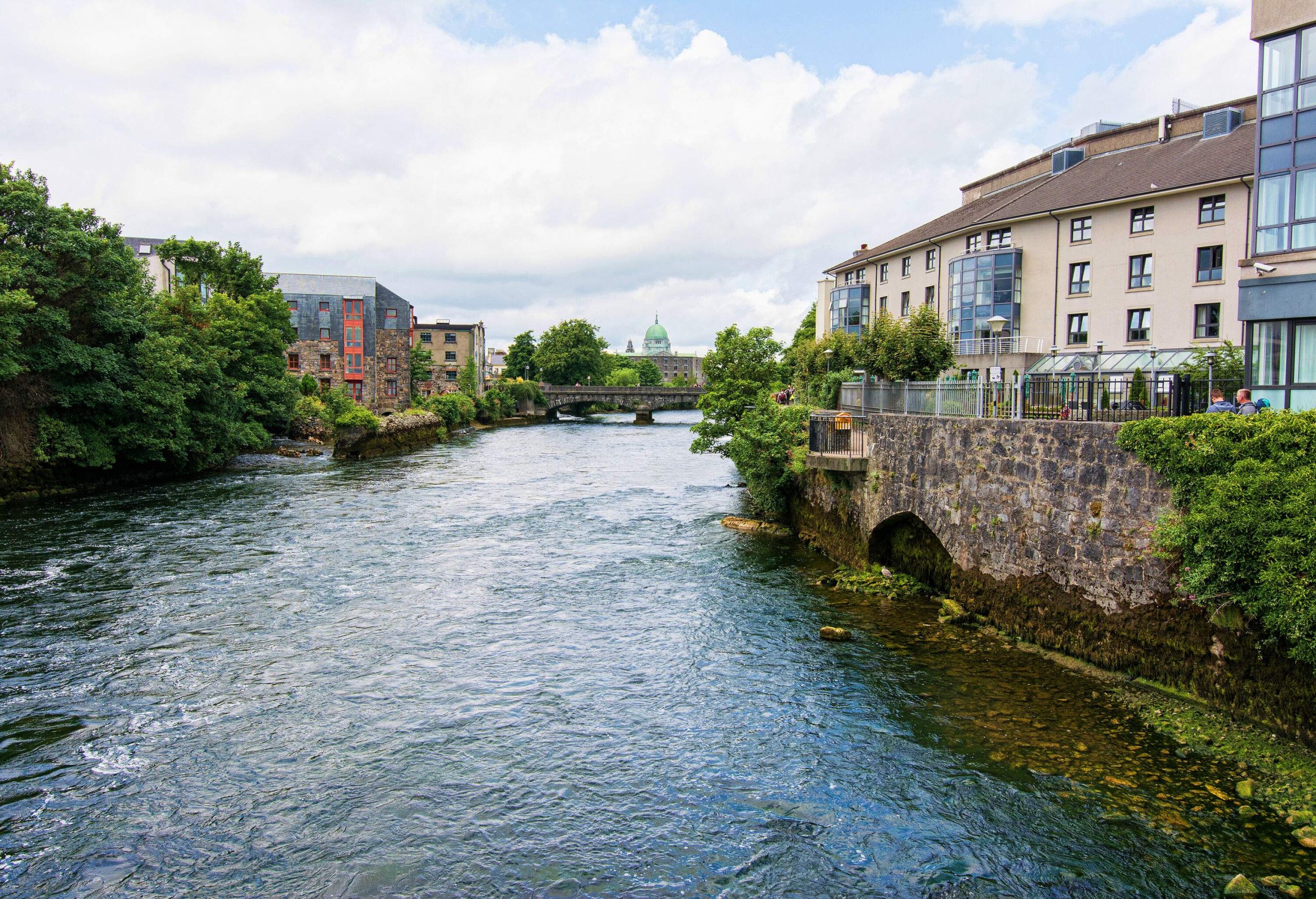 A mossy river in the centre of the riverside town with classic buildings against the cloudy blue sky.