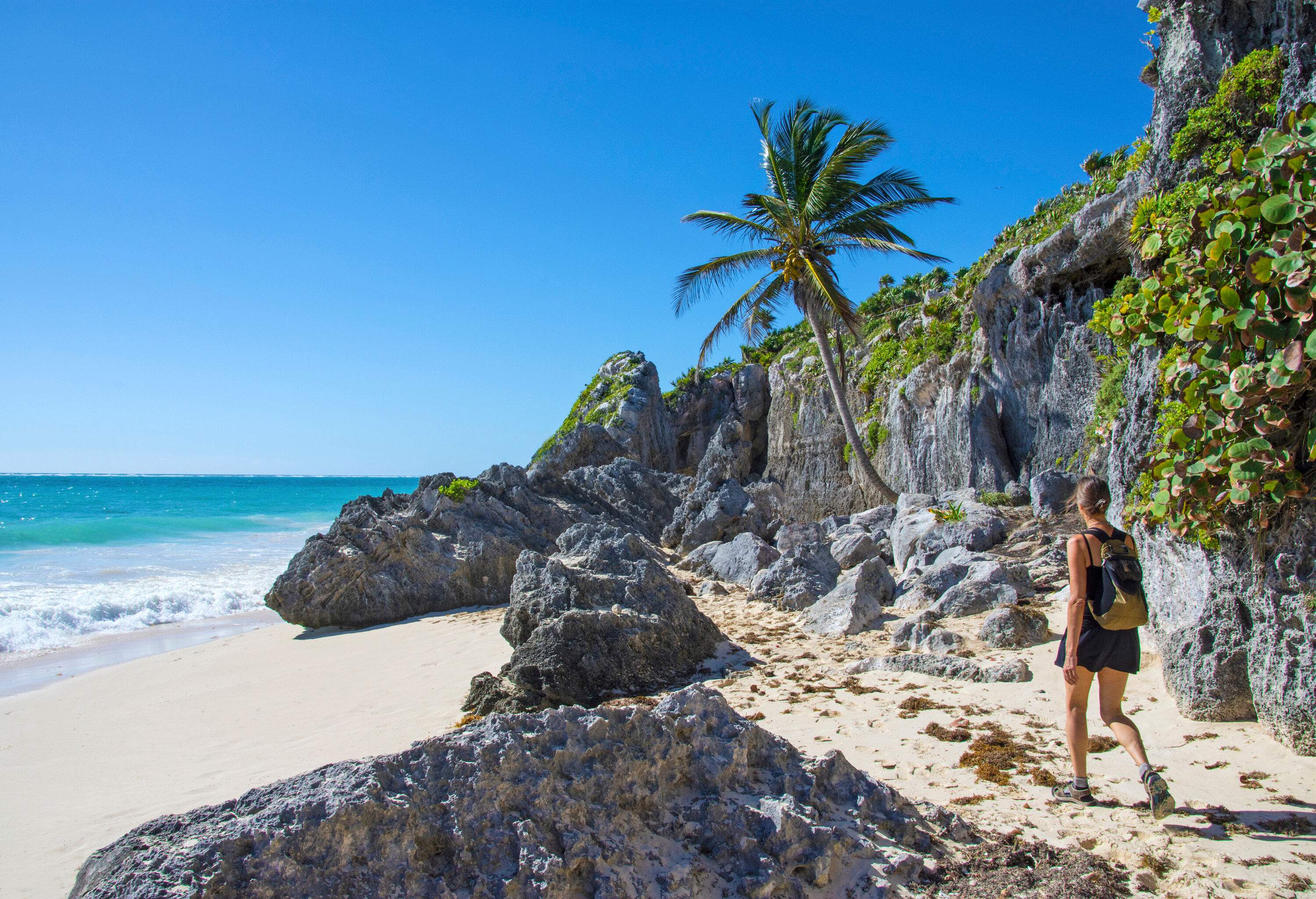 A woman walking along the rocky shore of a white sand beach.