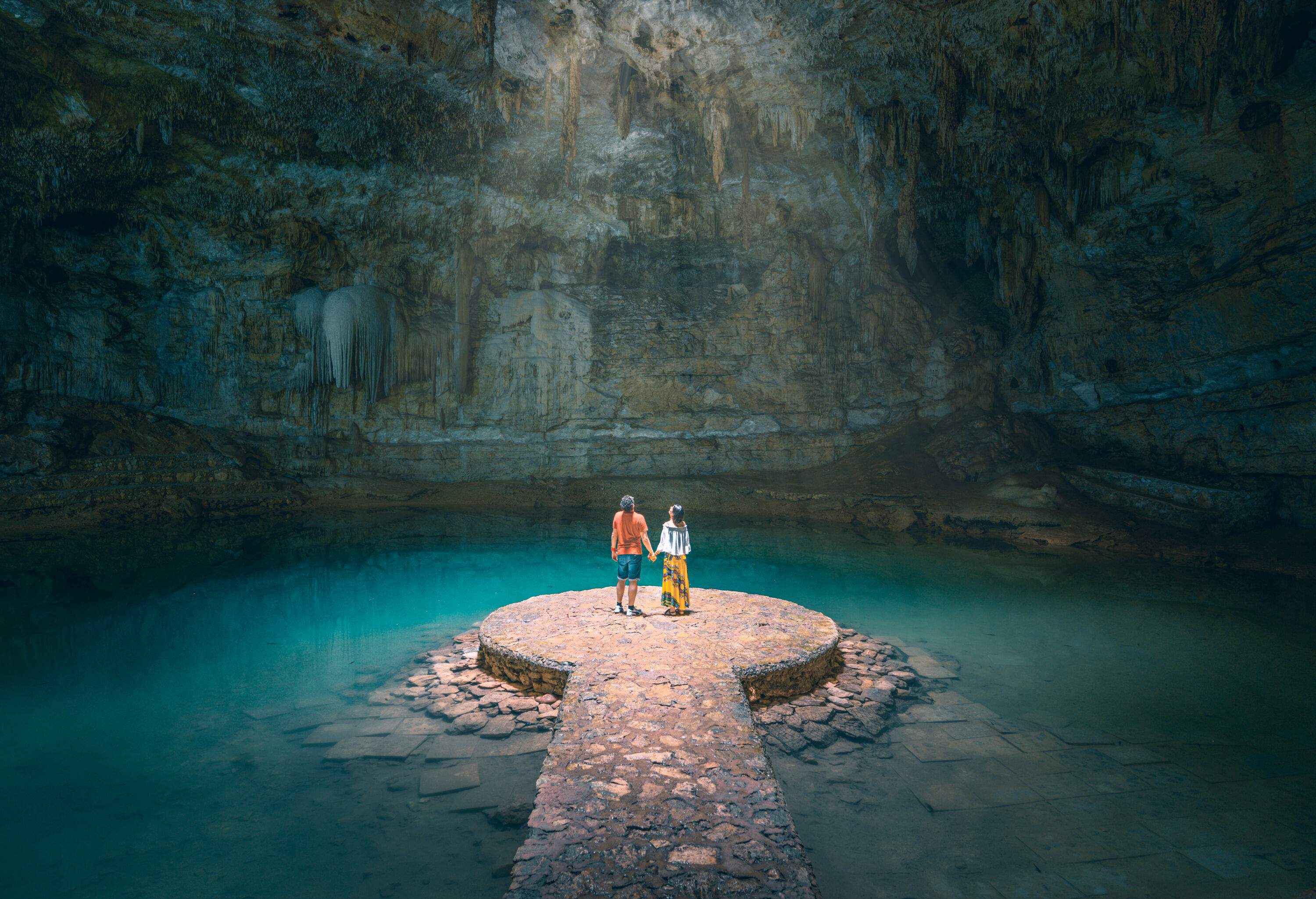 A holding hands couple looks up at the hole where the light passes through while standing on the rock platform.