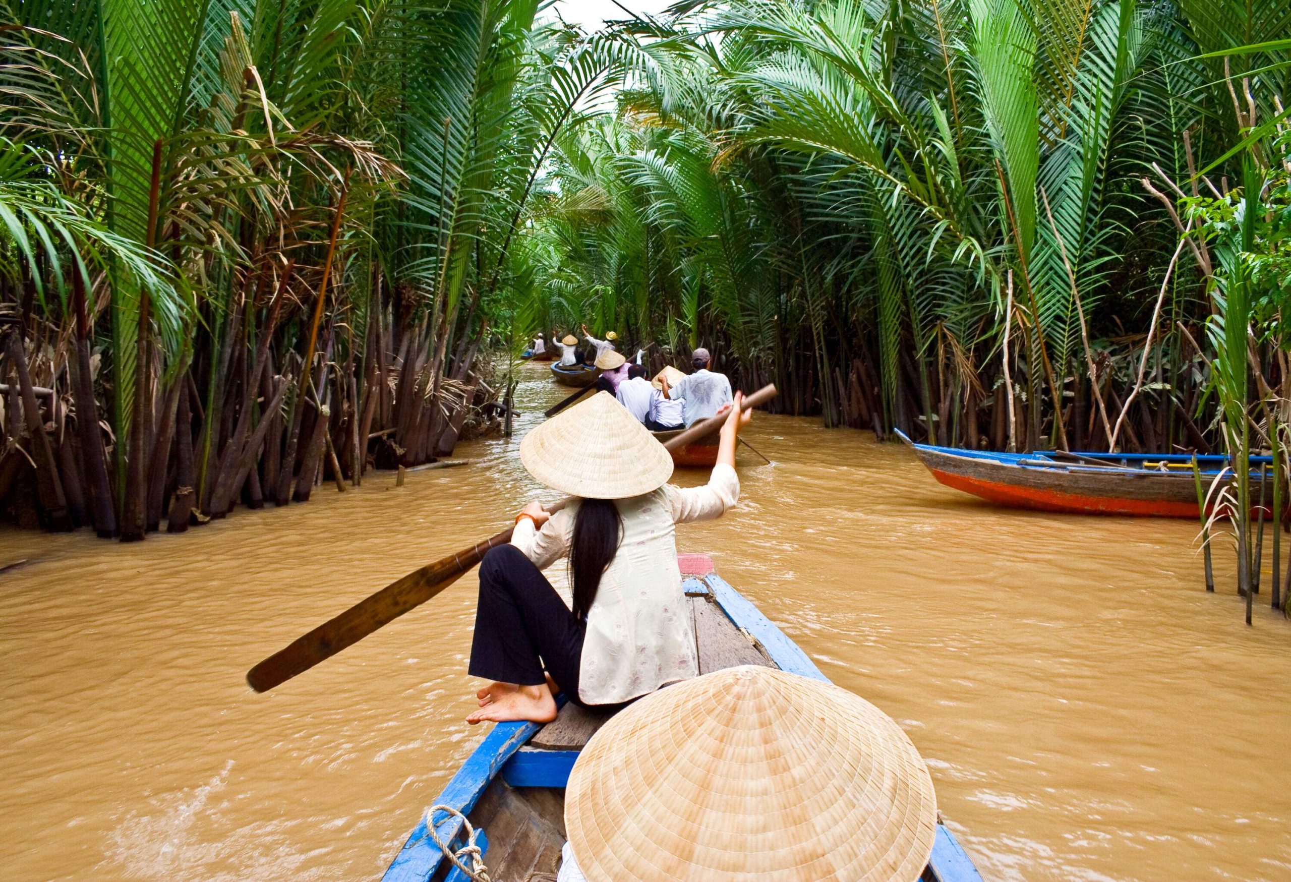 Individuals leisurely paddle on their wooden boats along a muddy river, enveloped by verdant foliage.