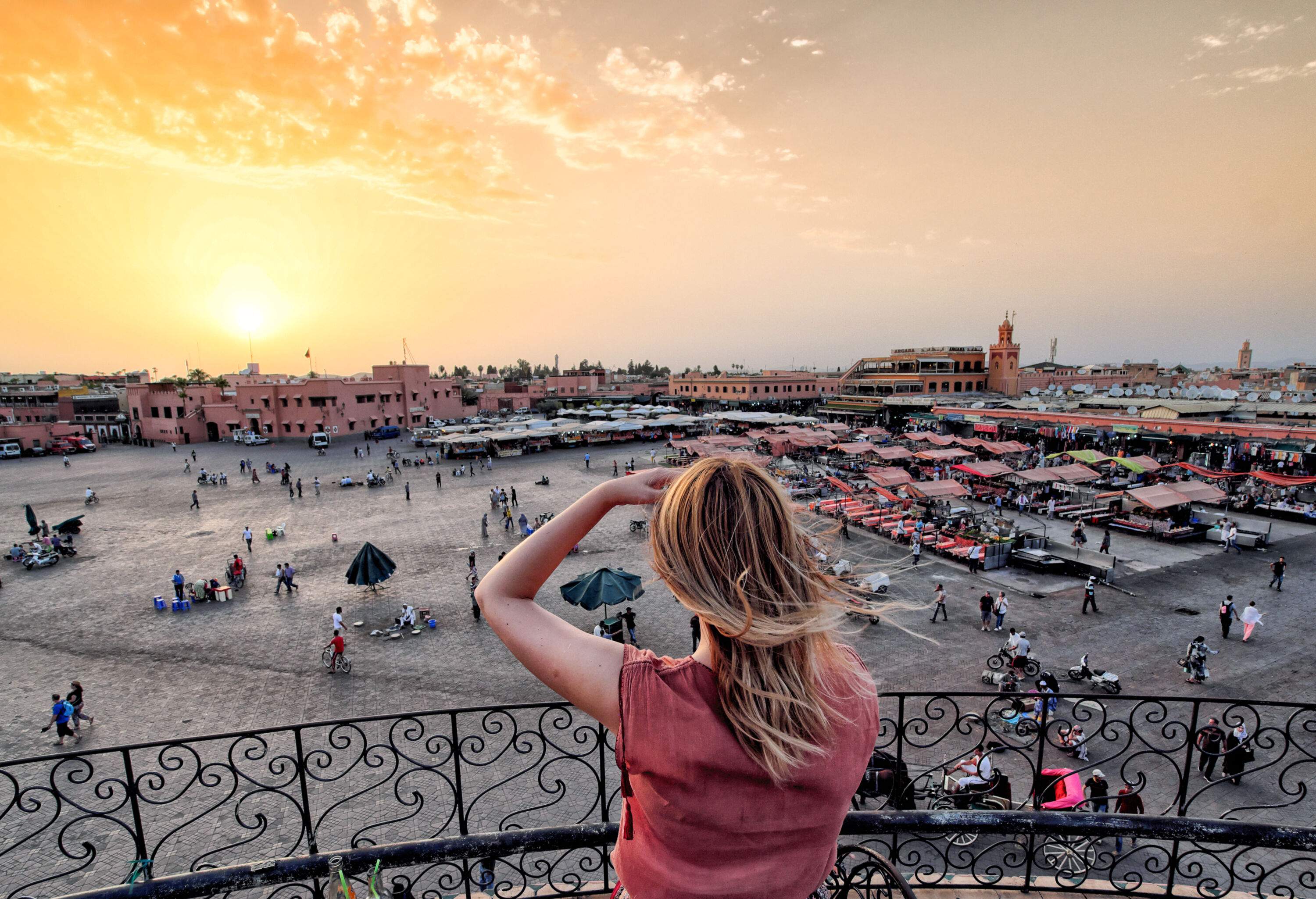 A woman stands on a terrace with overlooking views of a crowded market in a public square under a beautiful sunset.