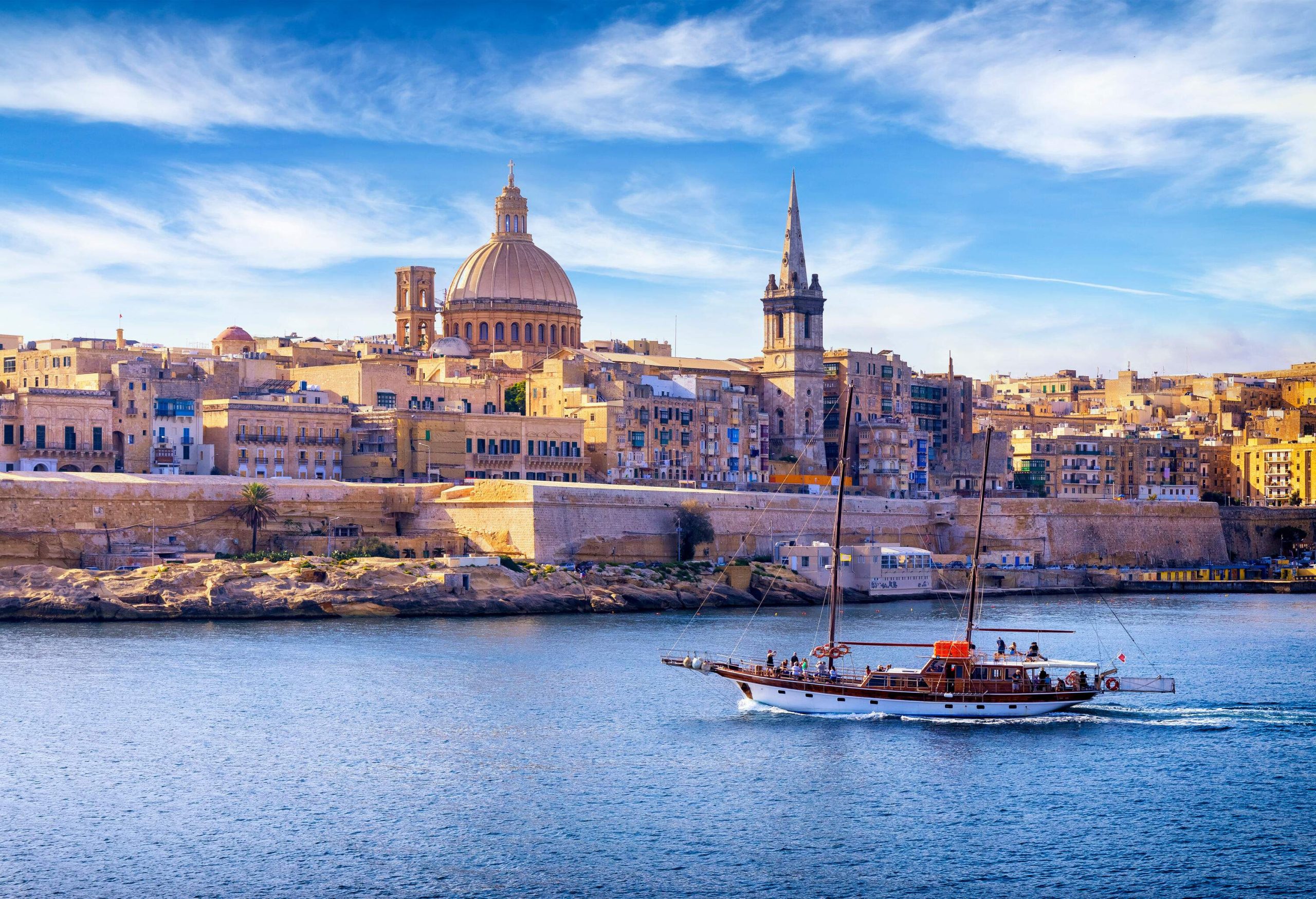 A dome cathedral with a pointed bell tower surrounded by city buildings overlooks a harbour with a cruising sailboat.