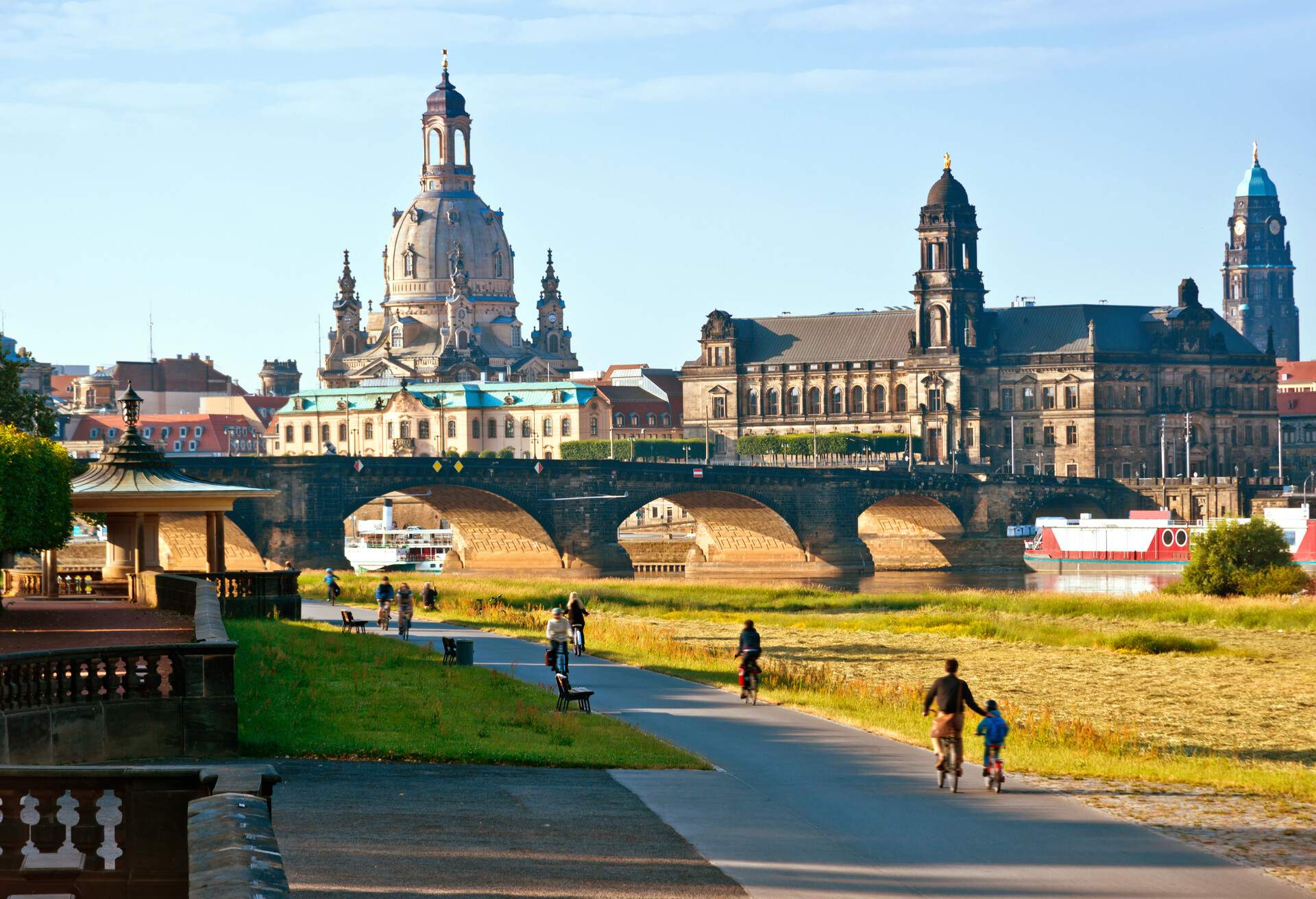 Several people bike in the cycle lane along a river spanned by an old arch bridge against the imposing visible towers protruding to the sky.