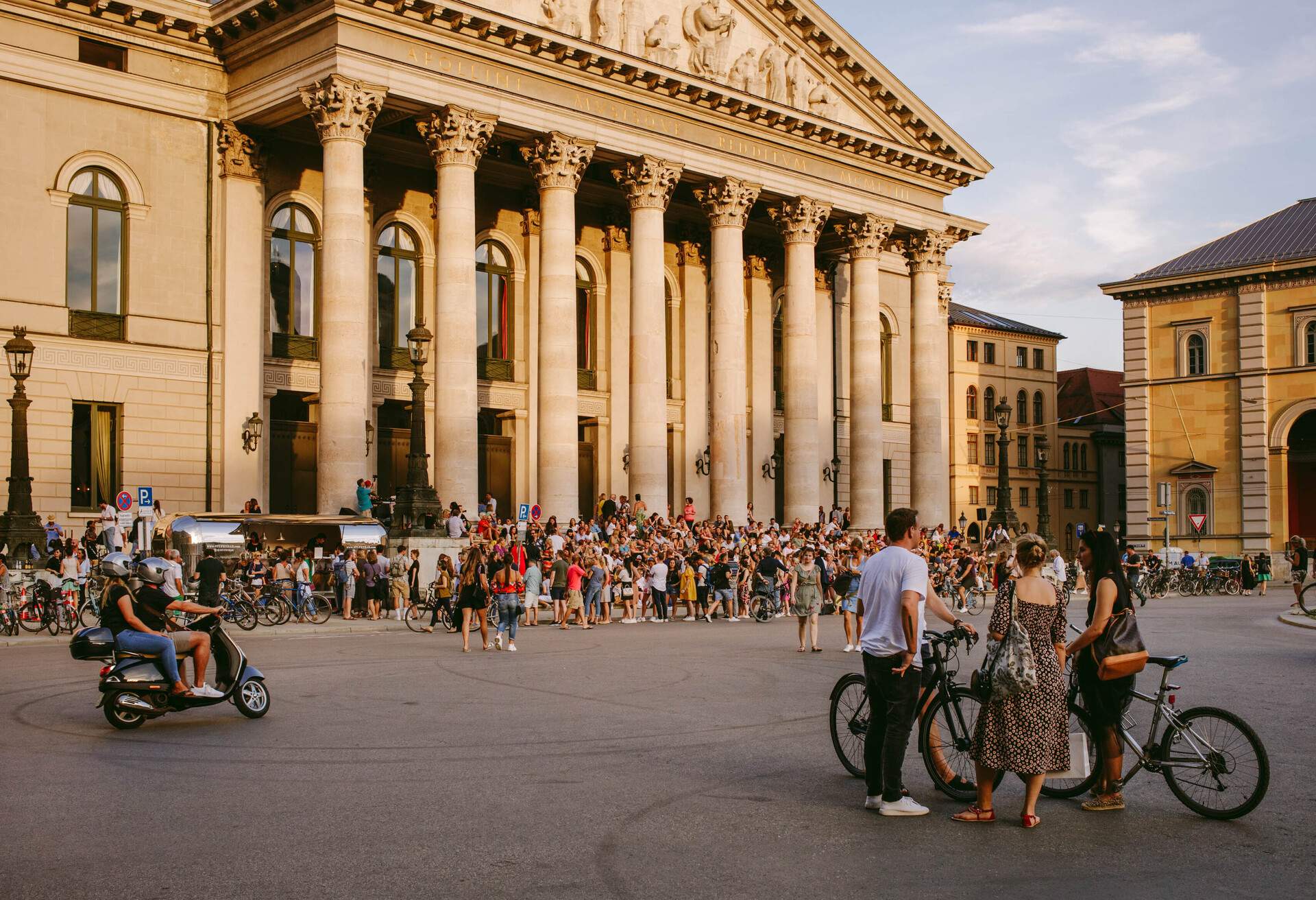 A bustling crowd seated outside a historic neoclassical-style theatre with pillars.