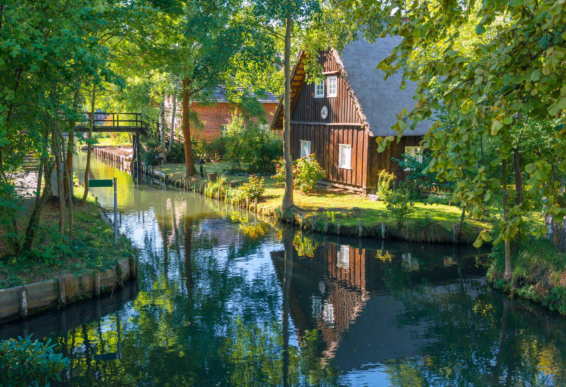 A cottage on the riverbank, adjacent to a wooden bridge, and surrounded by woods.