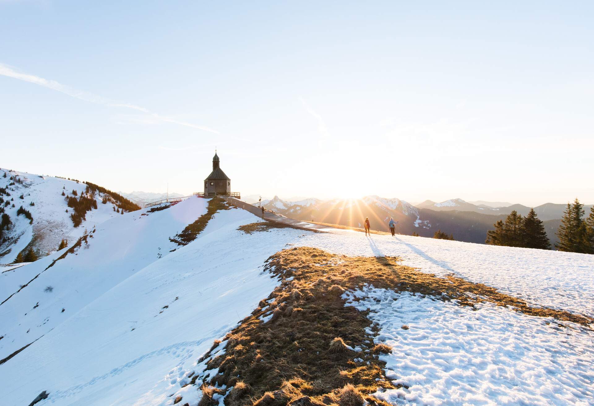 A small chapel on top of a mountain covered in snow against the clear blue sky and the beaming sun rays above a mountain range.