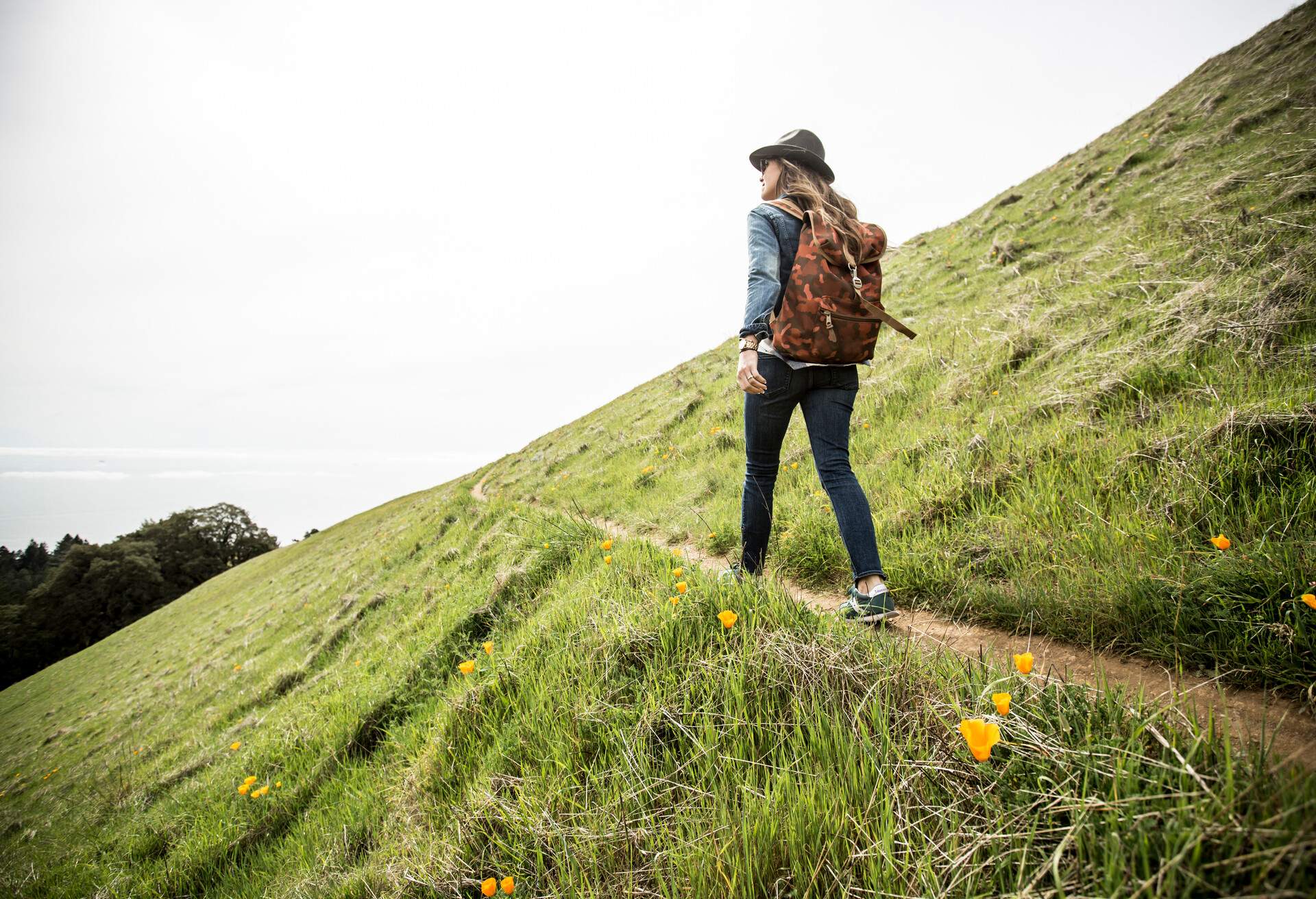 A woman carrying a backpack walking on a narrow trail on the hillside.