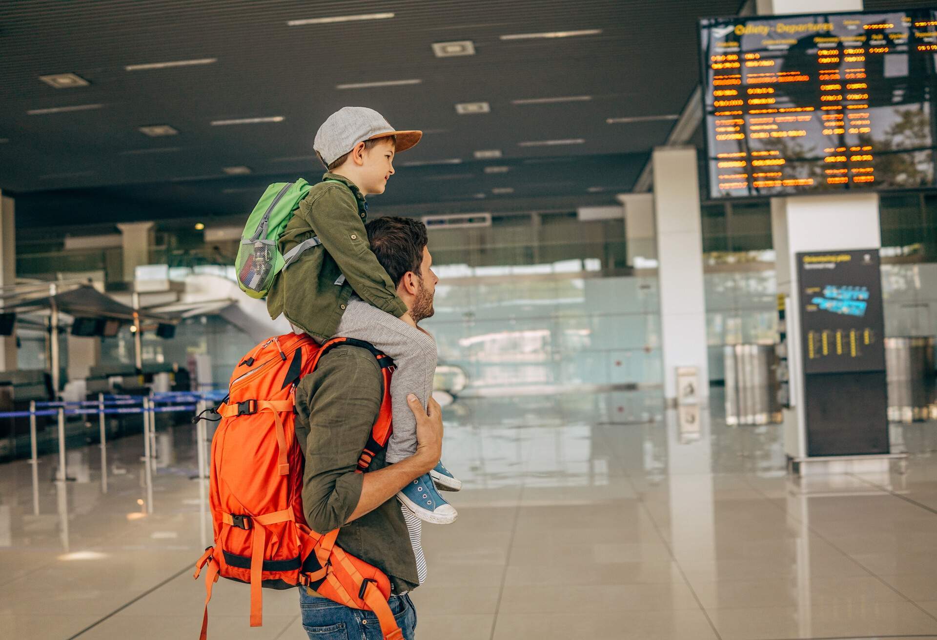 A man with an orange bag carries a kid on his shoulders while standing on an airport.