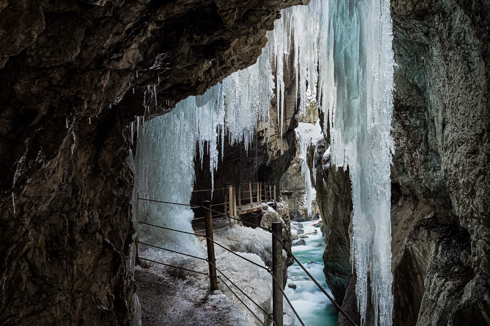 A fenced walkway on the sides of a gorge with massive icicles dangling above a flowing stream.
