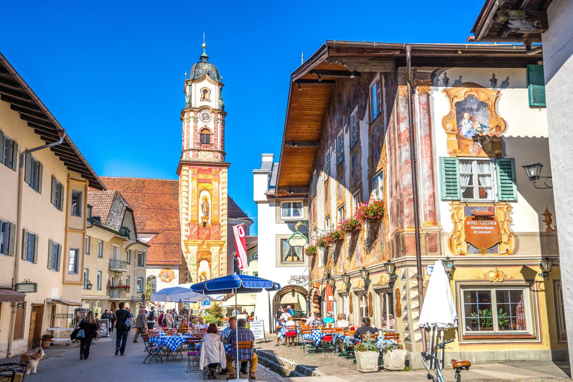 A crowded street with outdoor dining areas surrounded by restaurants and shops and a view of a church's belfry.