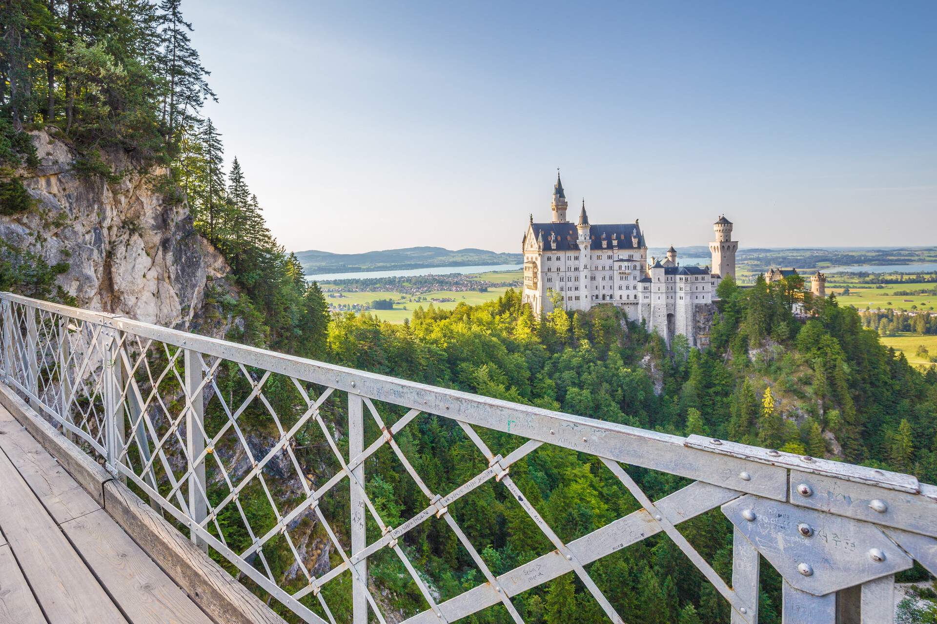 The Romanesque Revival palace of the famous Neuschwanstein Castle is surrounded by lush forest seen from the bridge.