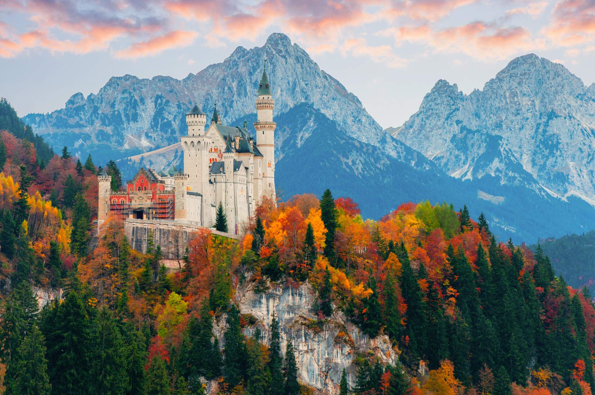 A medieval white castle with blue turrets on a rugged hill surrounded by trees with vivid autumn colours and a snow-capped mountain range in the background.