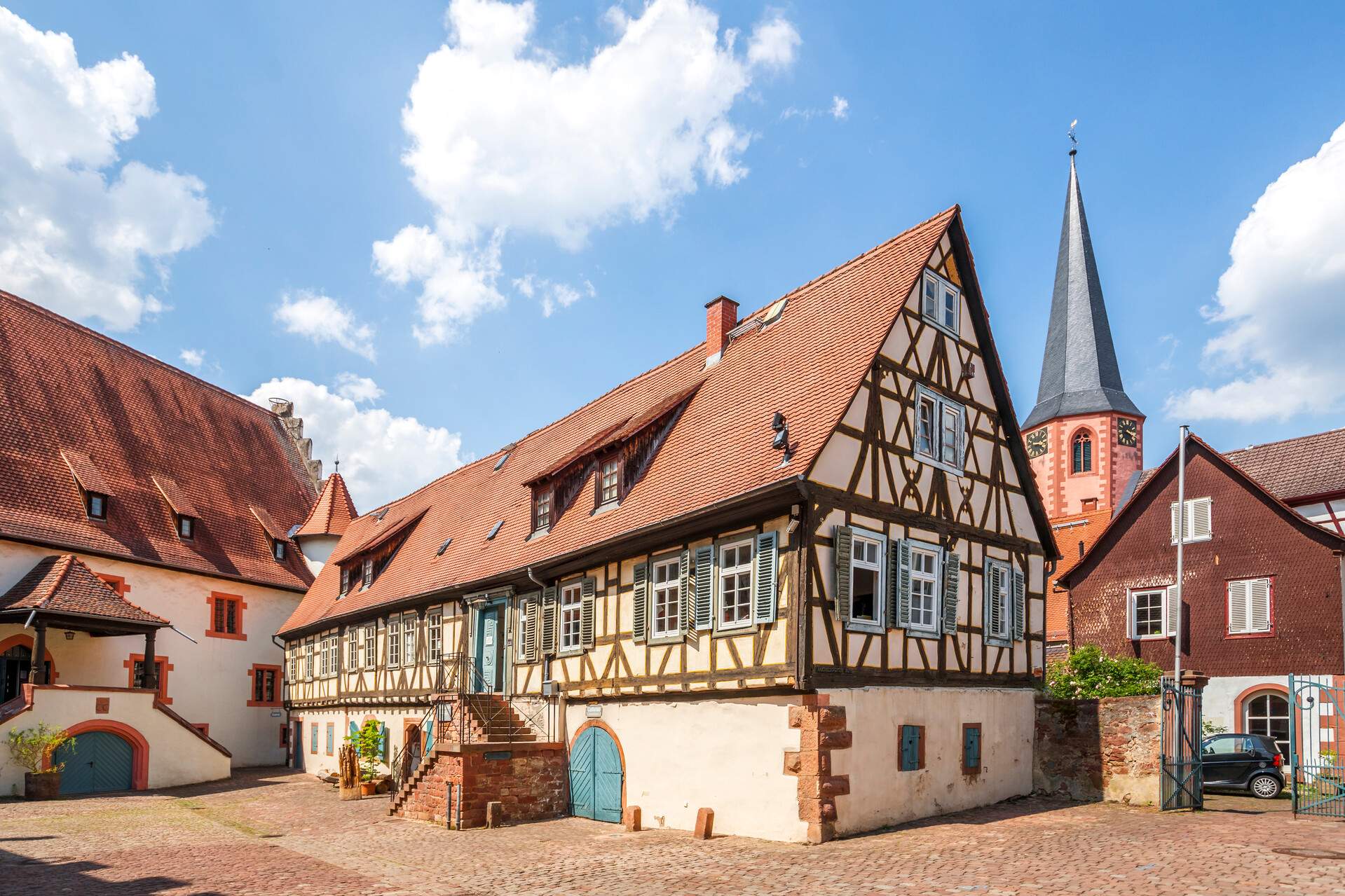 A large half-timbered home on a brick-paved street, backed by a tower with a tall, pointed roof.