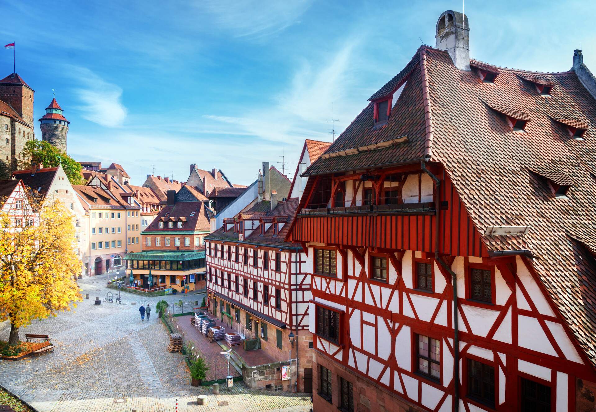 Two men walk on the cobbled street alongside lined tall colourful half-timbered buildings.