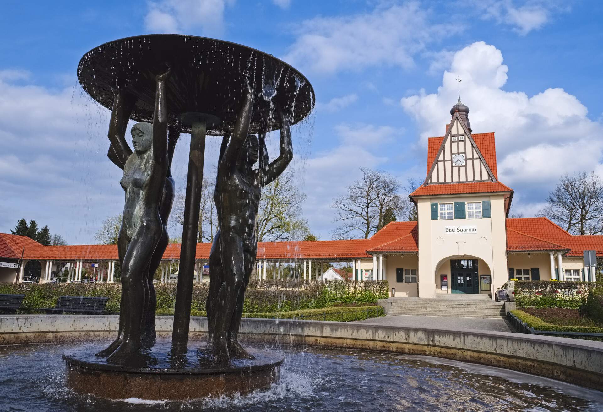 A fountain with black sculptures of people lifting a circular dish near the entrance to a white train station.