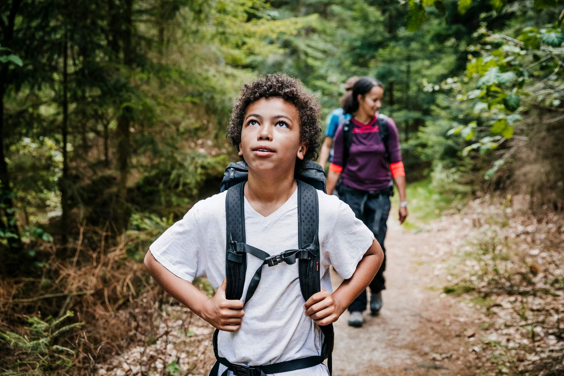 Young kid in white shirt carry a bag and looks up while hiking in the forest with people following behind him.