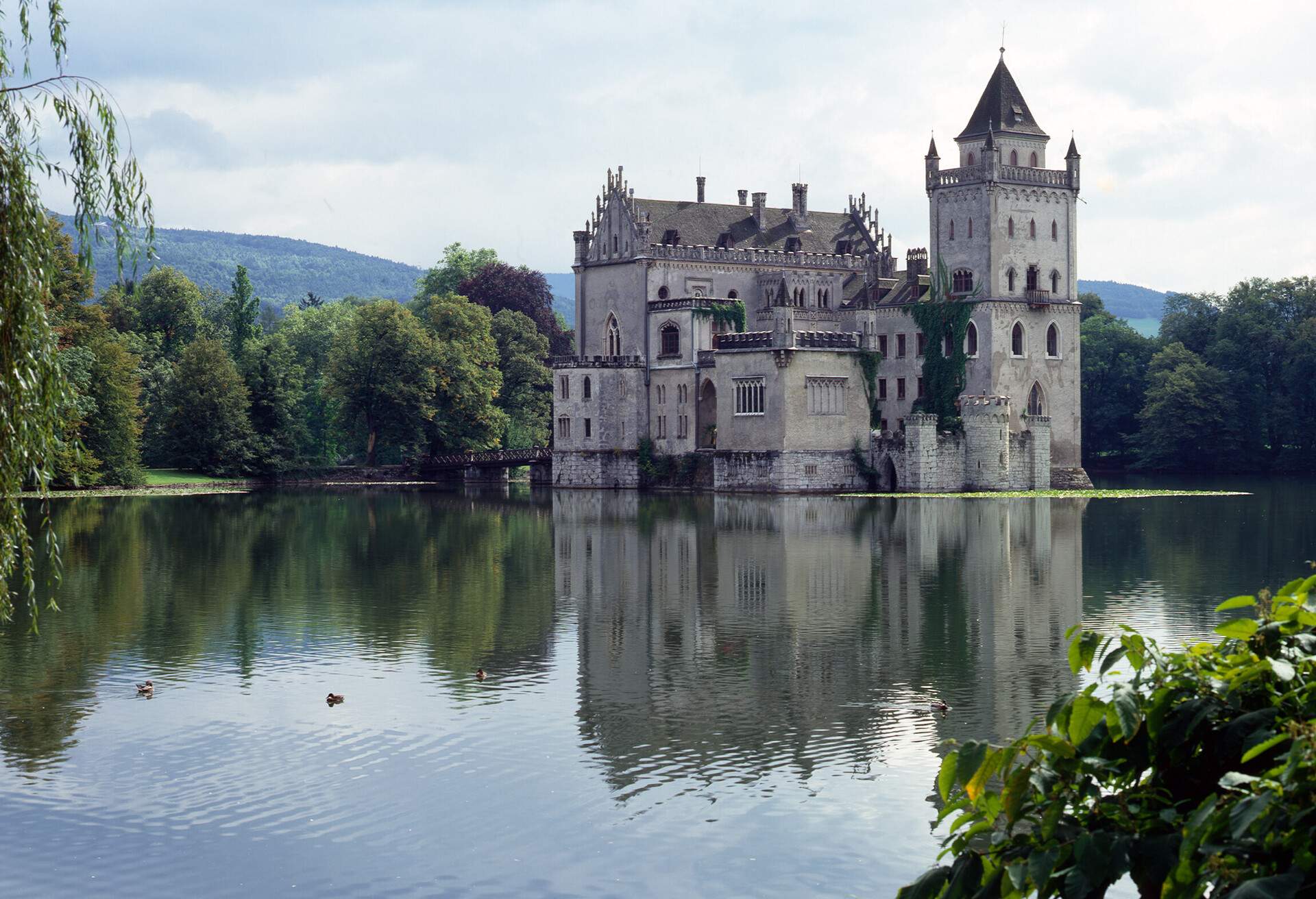 A low-lying water castle surrounded by a forest of trees and distant mountains.