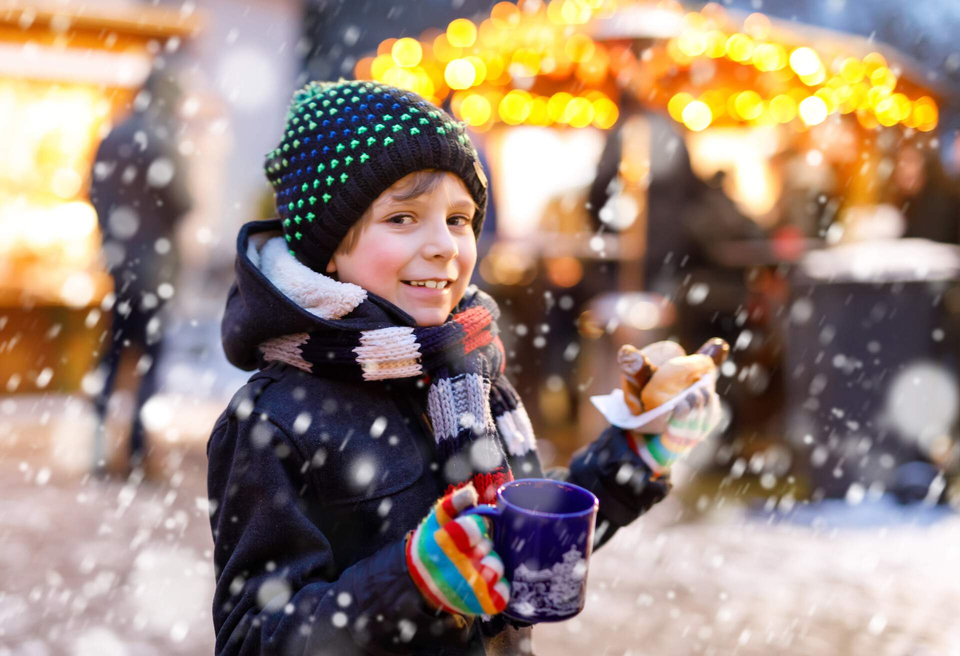 Little cute kid boy eating German sausage and drinking hot children punch on Christmas market. Happy child on traditional family market in German. Laughing boy in colorful winter clothes.