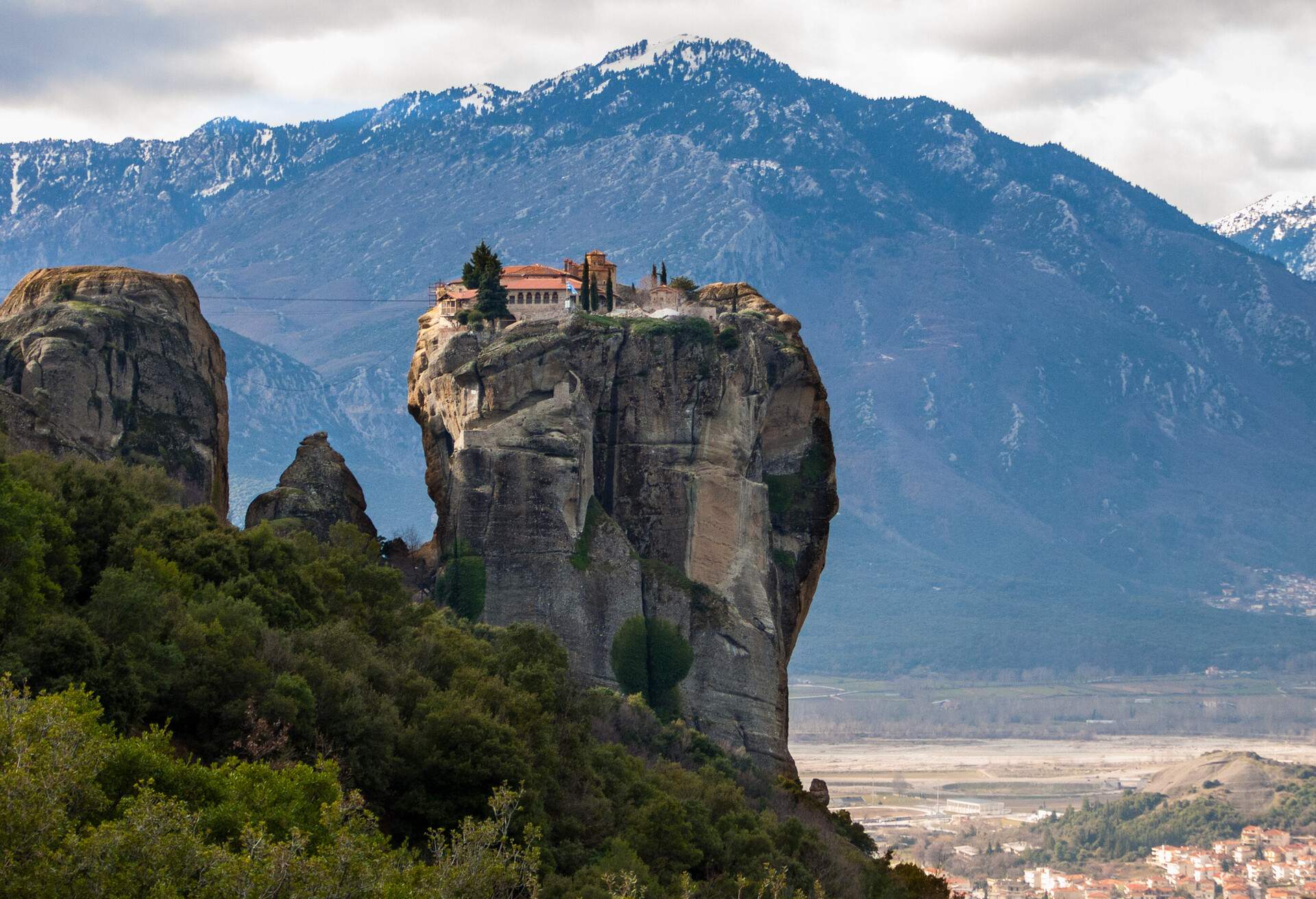 A monastery on top of a steep rock formation against the backdrop of snow-capped mountains.