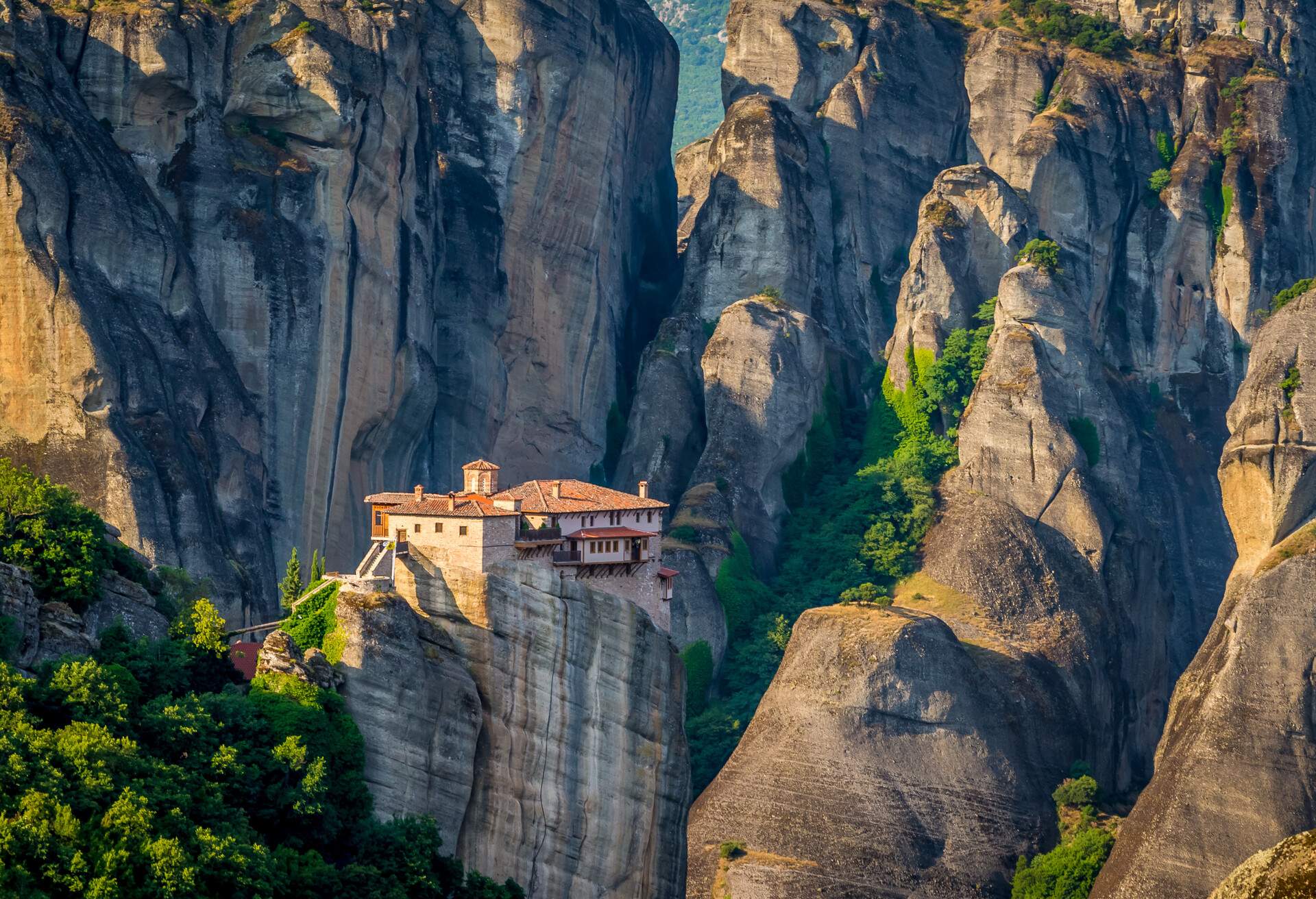 A majestic monastery sits atop a steep rock formation, set against a stunning backdrop of tall, sheer rock cliffs.