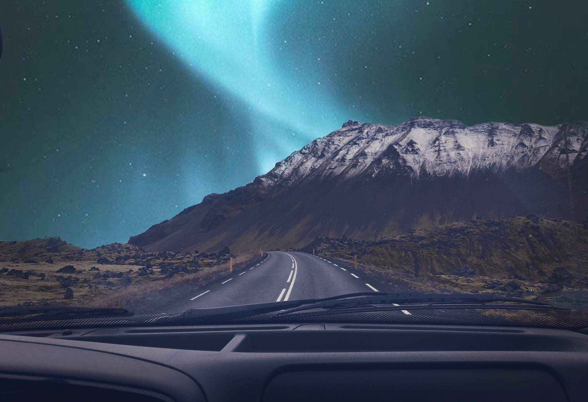 A car driving down the road as the northern lights swirl around a snow-capped mountain in the distance.